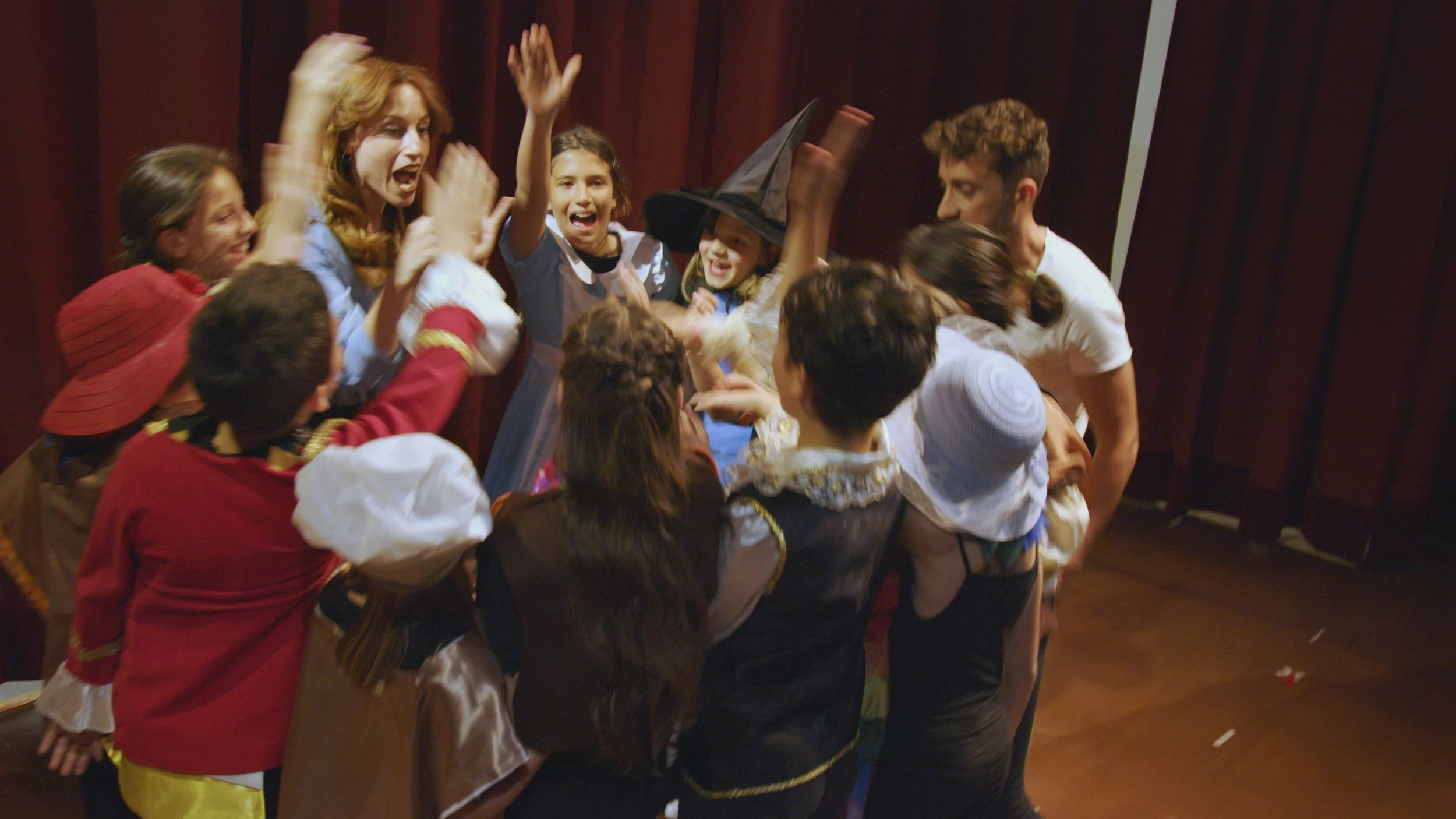 Gruppo di bambini e adulti in costume che festeggiano e si abbracciano, con un cappello da strega tra loro, in un ambiente con tende rosse sullo sfondo.