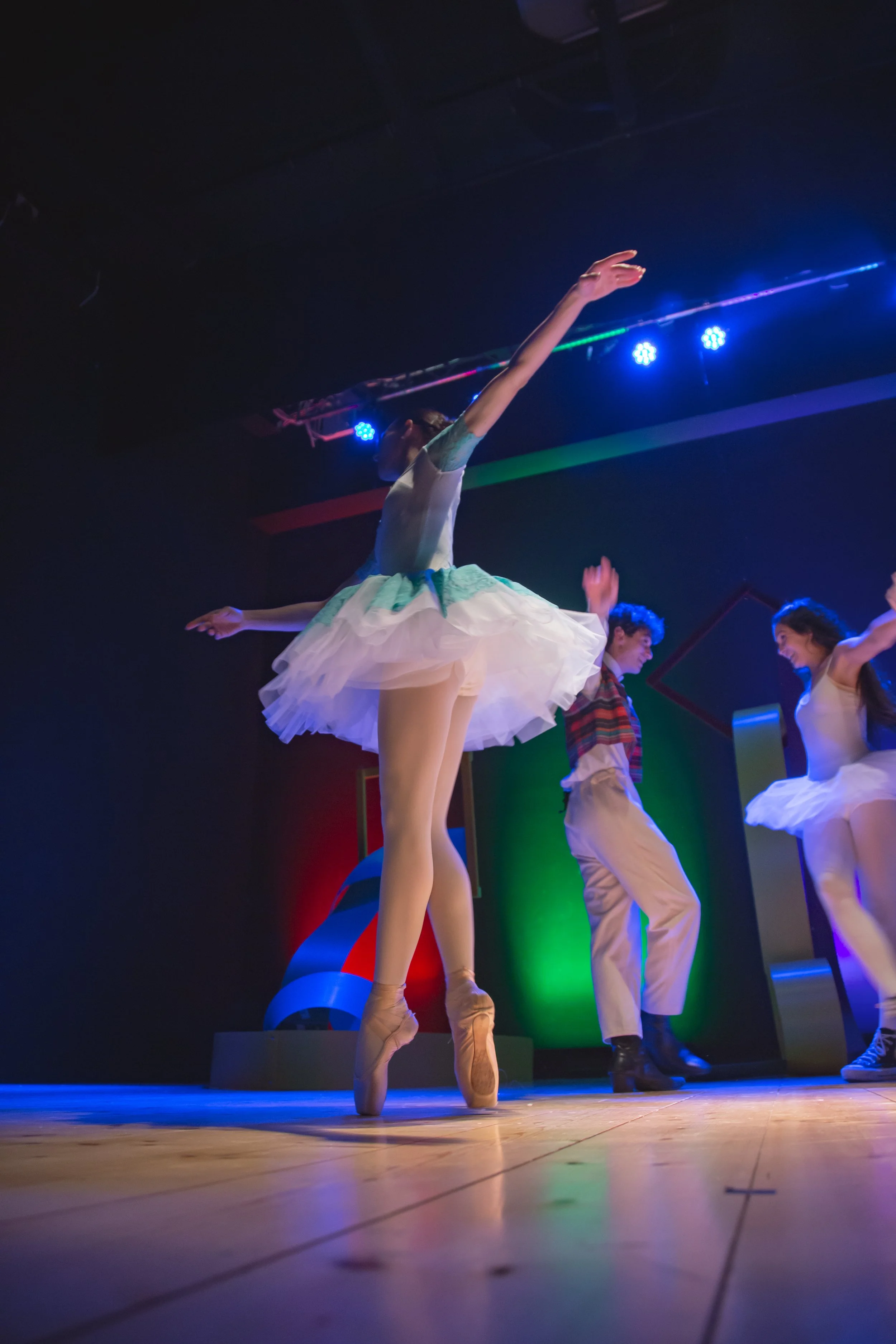 Ballerina in tutù rosa che danza sul palco con altre persone sullo sfondo, illuminata da luci colorate in un teatro.