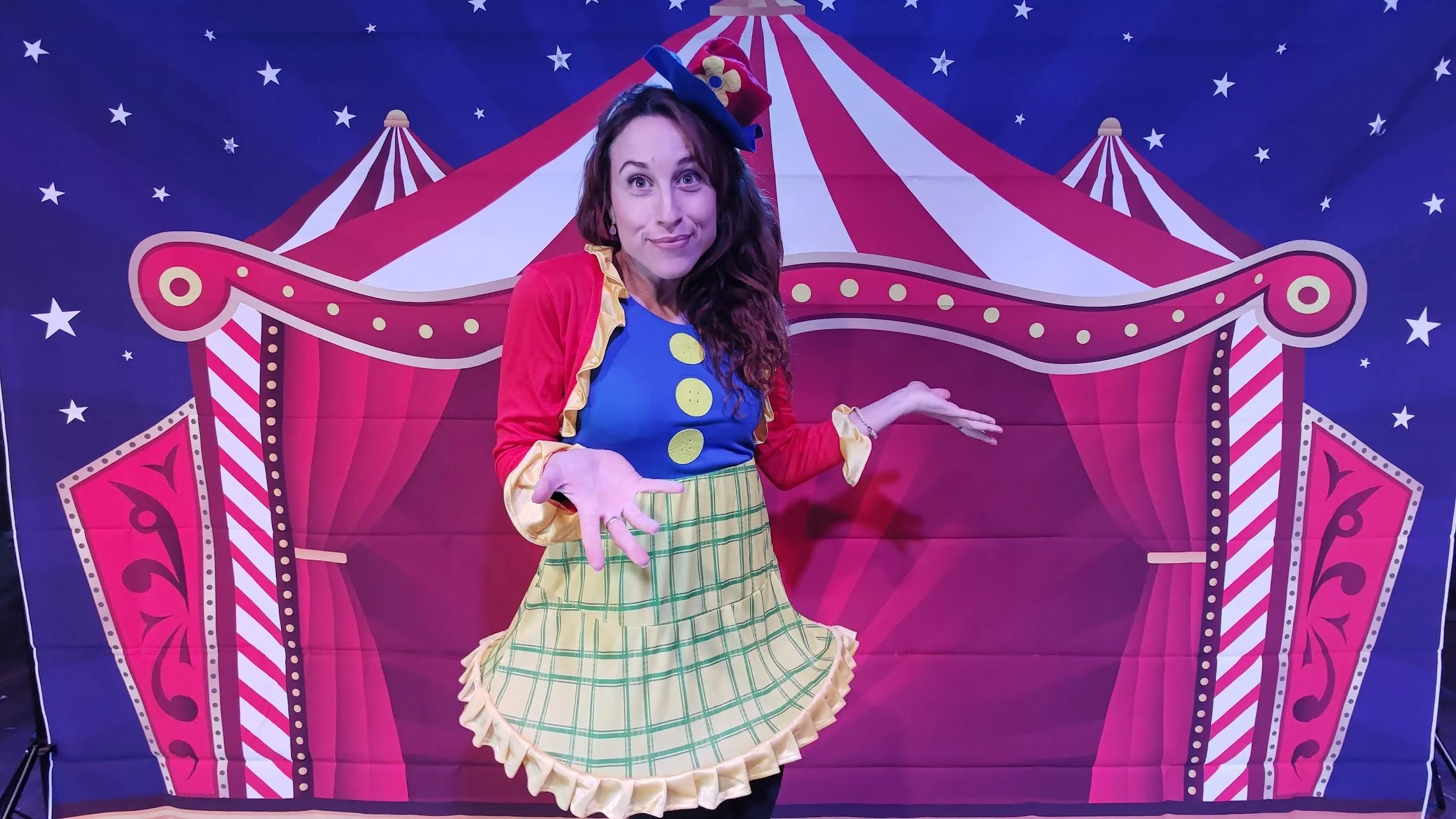 Una donna travestita da clown davanti a uno sfondo di un tendone da circo rosso e bianco con stelle blu, sorridente con braccia aperte e cappello a cilindro colorato.