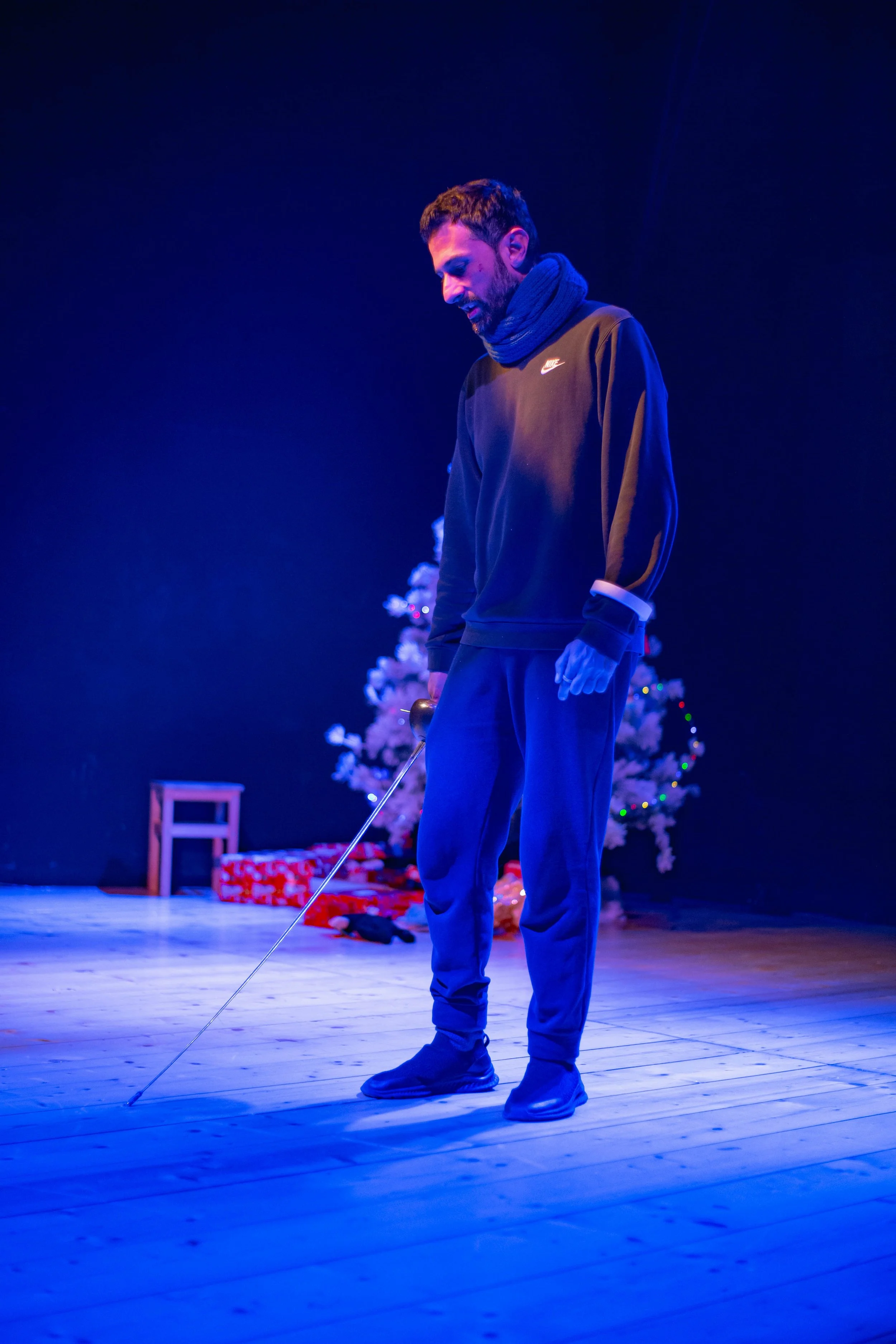 Uomo con cappuccio e sciarpa che tiene una canna da golf in un ambiente decorato per Natale, con un albero di Natale e regali sullo sfondo, illuminato da luci blu.