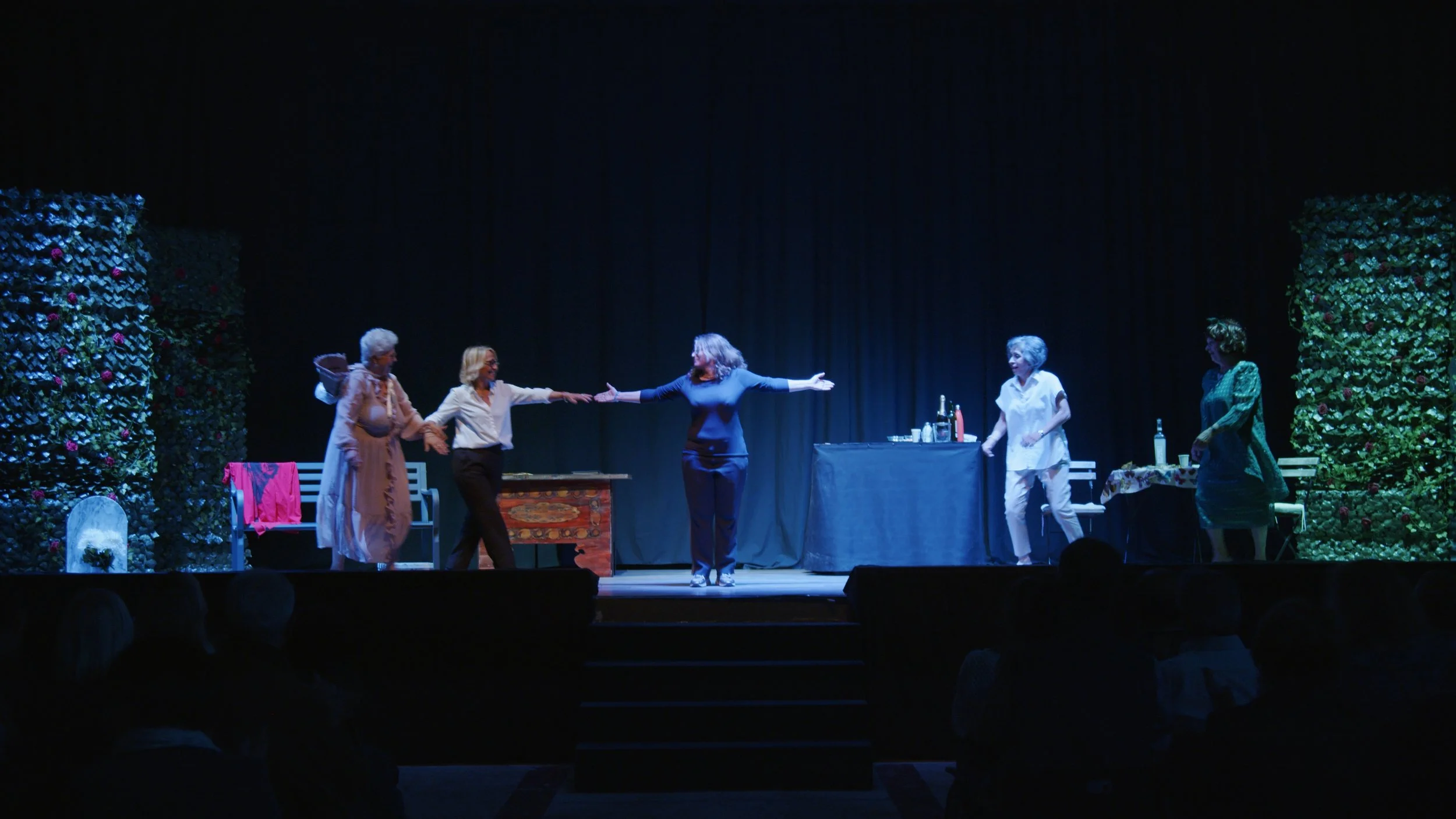 Scene teatrale con cinque donne che interagiscono, alcune in piedi, con decorazioni floreali e un tavolo con bottiglie e utensili, sorretta da un palco teatrale con sfondo scuro.