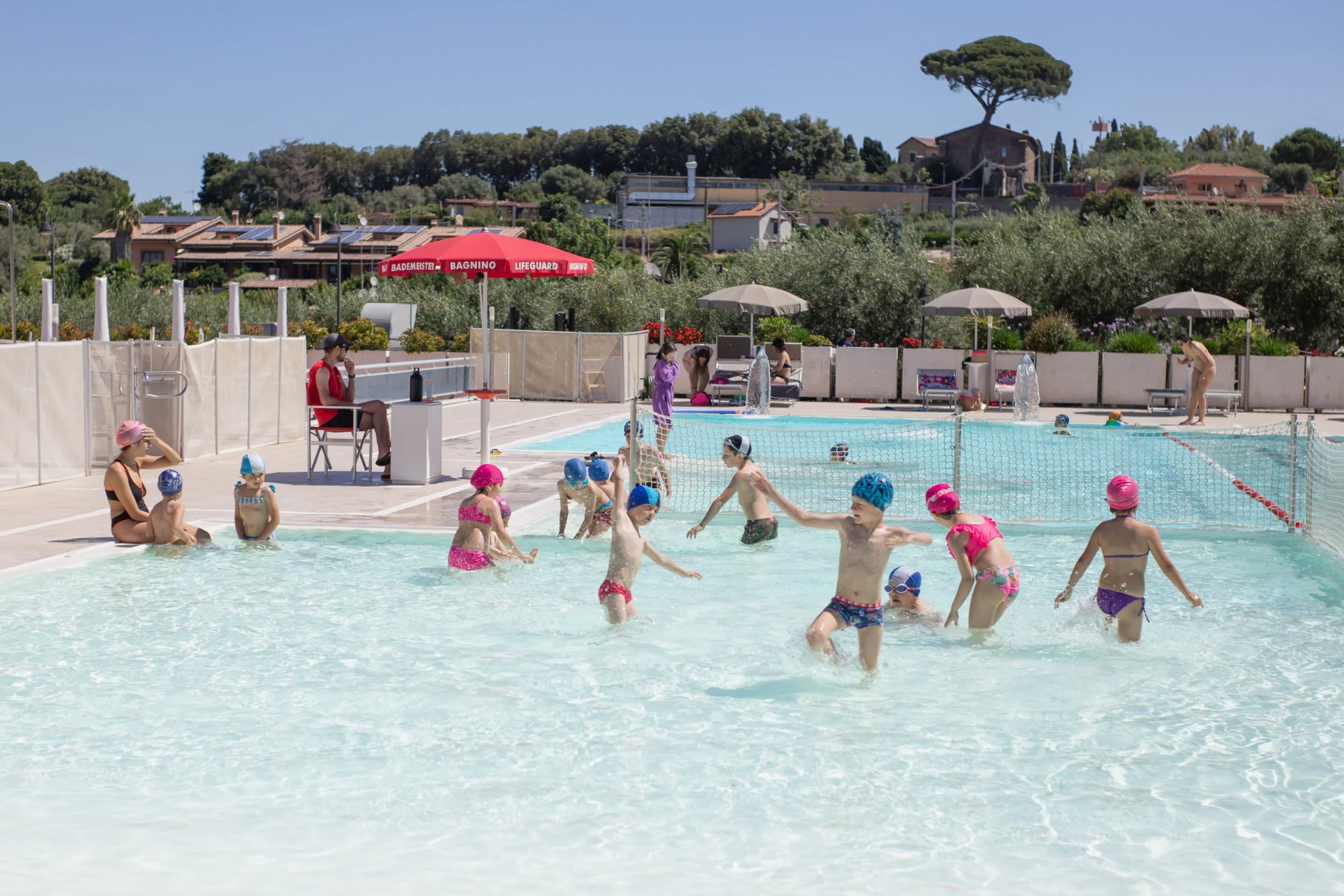 Bambini che giocano e nuotano nella piscina all'aperto, con sedie, ombrelloni e un paesaggio di case e alberi sullo sfondo.