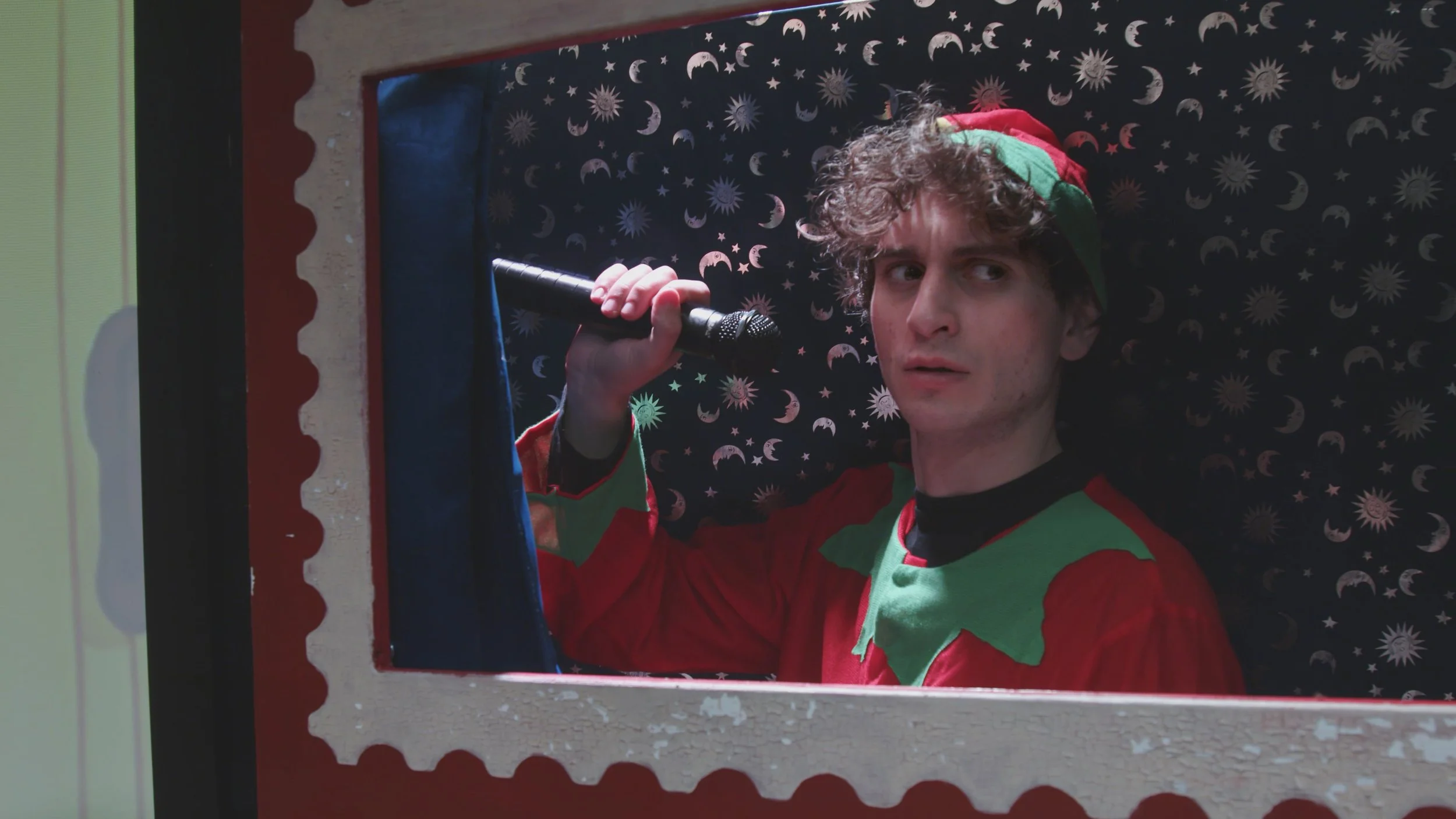 Ragazzo vestito da elfo con cappello rosso e verde, che guarda dalla finestra decorata con tema natalizio, con sfondo a stelle e lune.