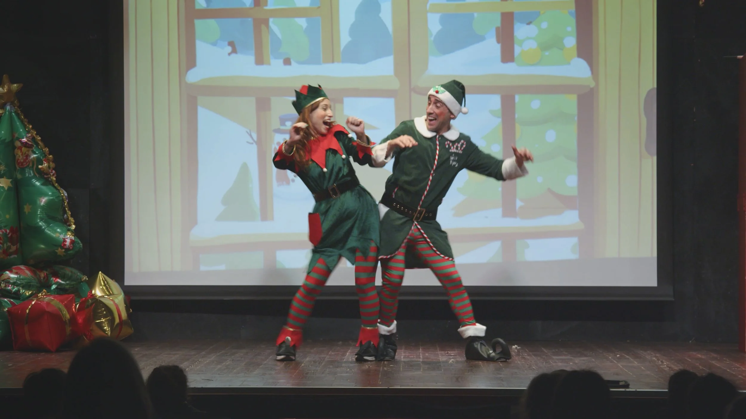 Due persone vestite da elfi di Natale che danzano sul palco con uno sfondo di un disegno natalizio e decorazioni, tra cui un albero di Natale con pacchi regalo sotto.