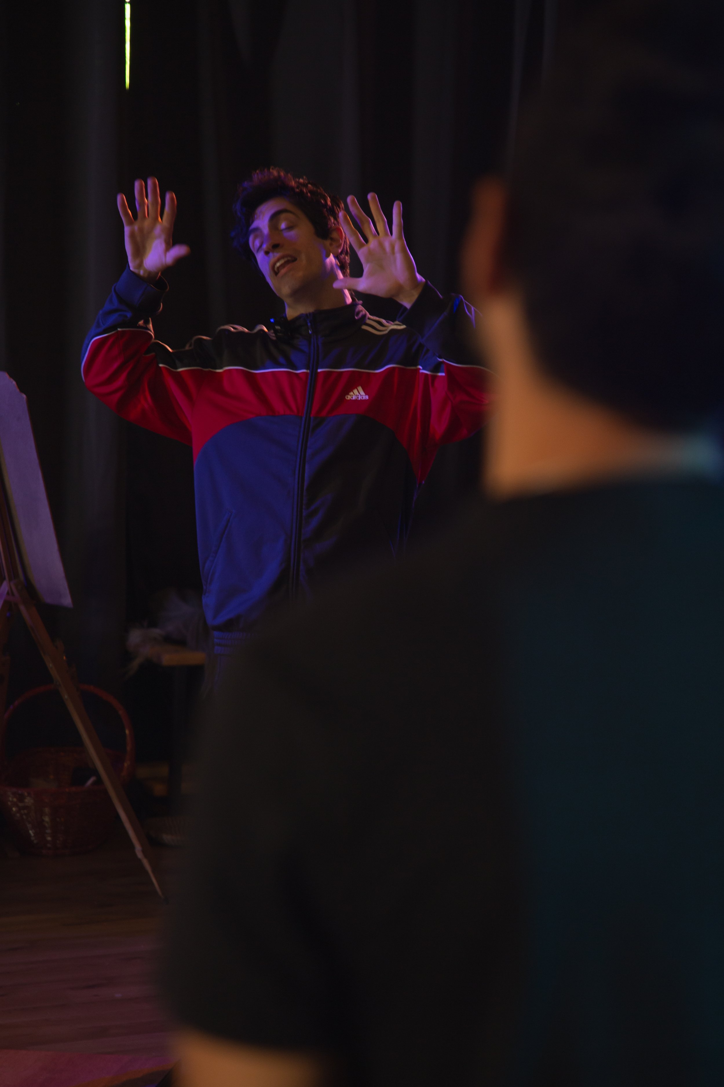 Un uomo con capelli scuri e sui vestiti rossi e blu, con le mani sollevate, sembra fare un'espressione di sorpresa o emozione, in un ambiente buio con luci soffuse.