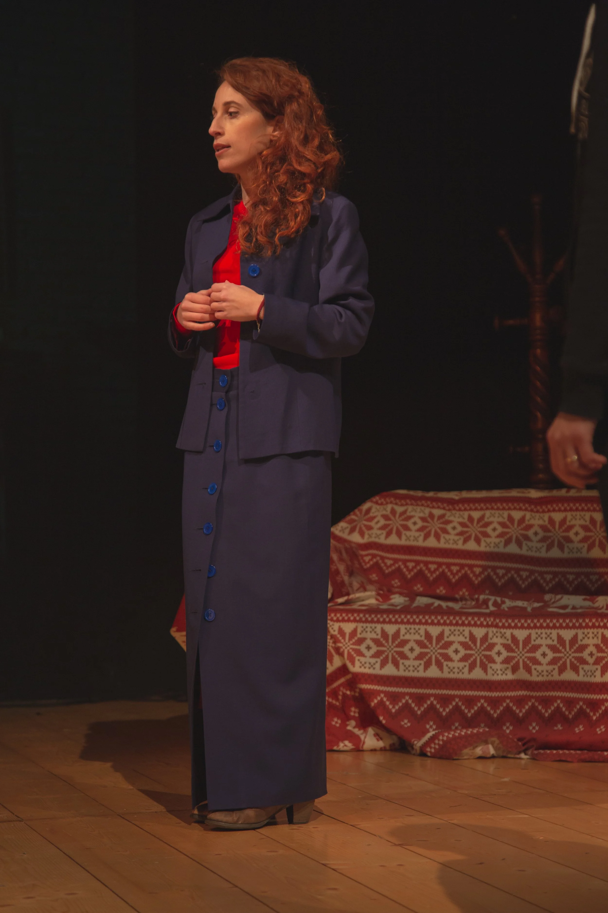 Una donna con capelli ricci rossi indossa un abito lungo blu e una giacca blu, sta in piedi sul palco con un palco di legno e una sedia a motivi natalizi sullo sfondo.