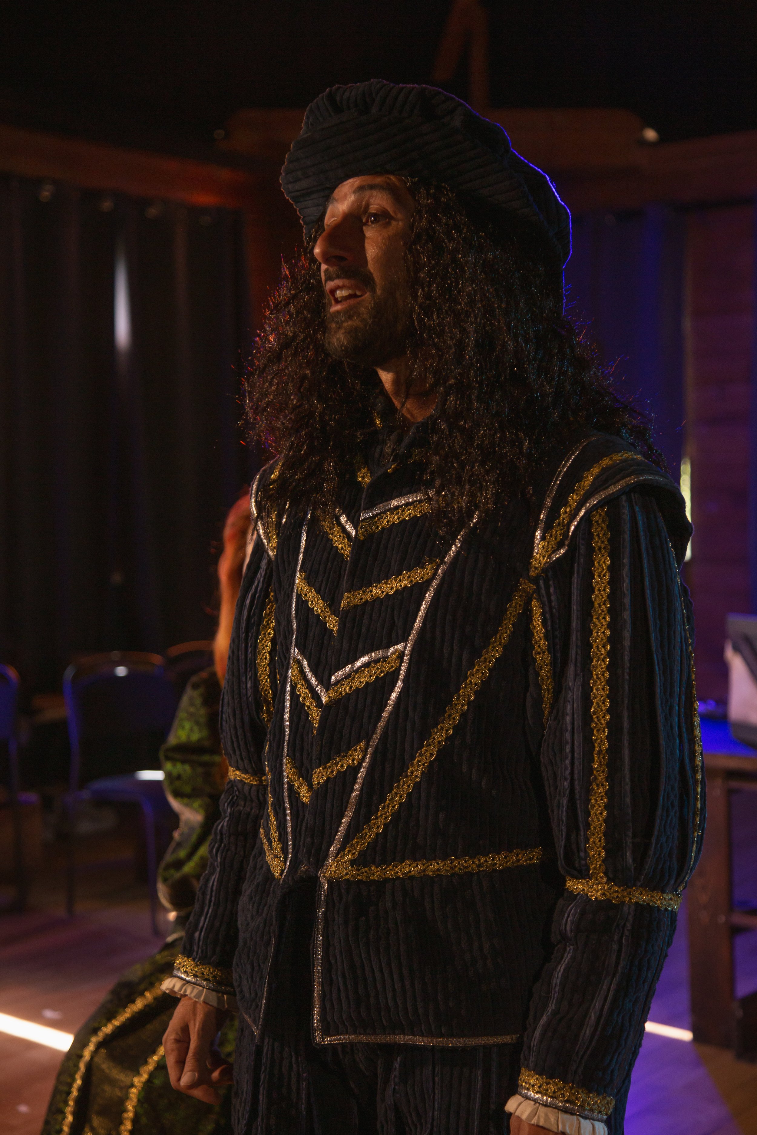 Persona vestita con abiti storici in velluto nero con dettagli dorati, cappello nero a tricorno, ambiente con luci soffuse e sfondo scuro.