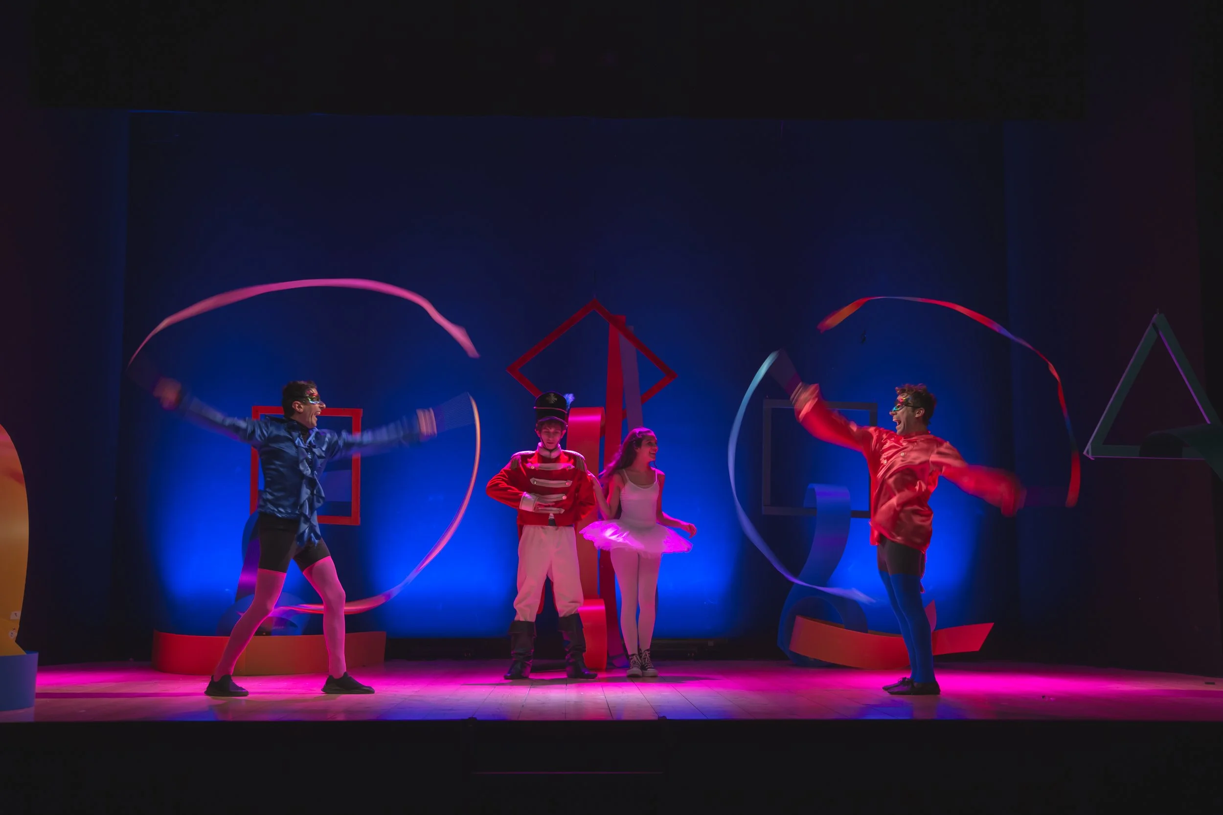 Performers in colorful costumes on stage with geometric decorations and ribbons, illuminated by stage lights.