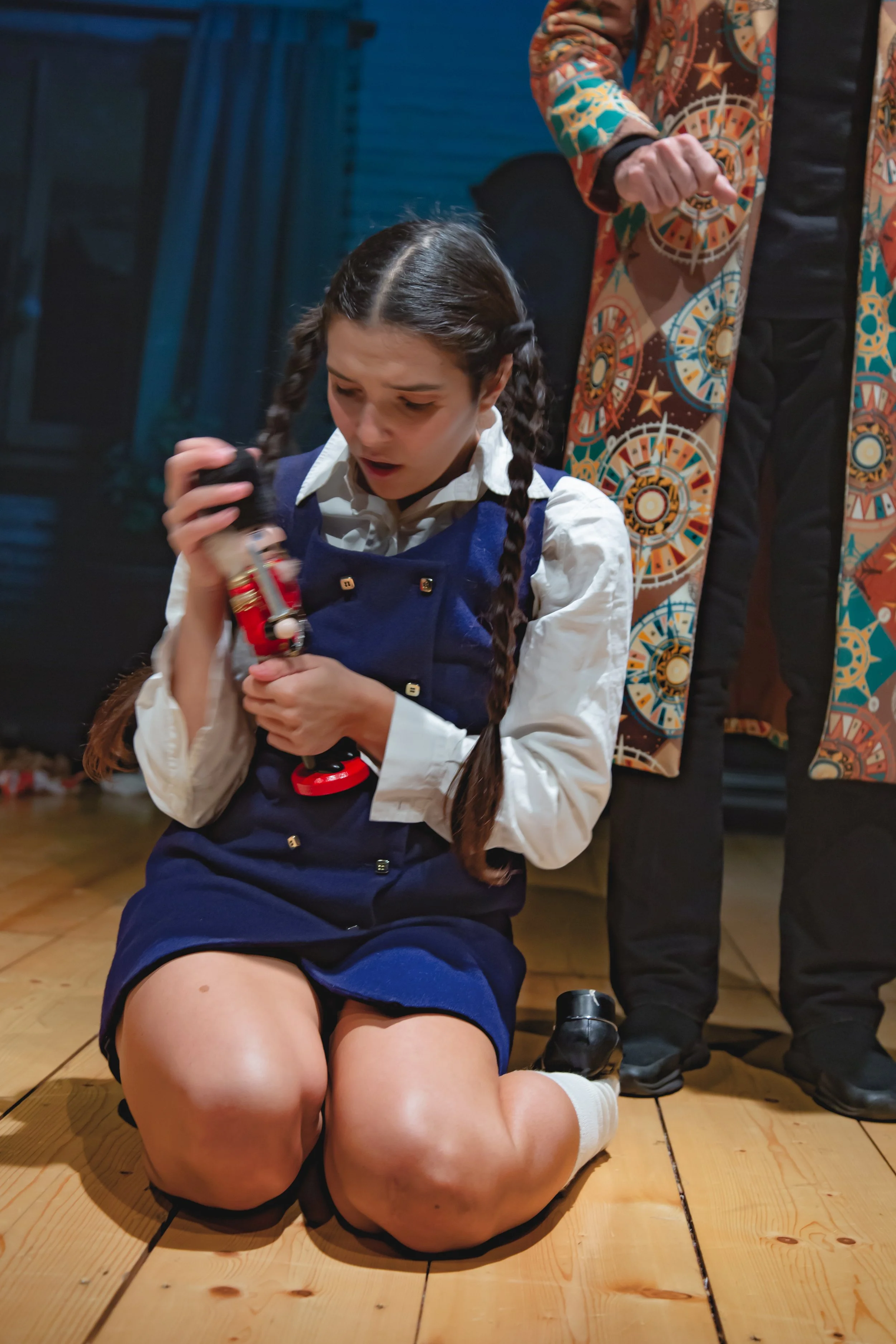 Una ragazza con treccine, vestita con una divisa scolastica blu, è seduta a terra con le gambe piegate, che tiene in mano una figura giocattolo. Dietro di lei, in piedi, c'è una persona con una giacca colorata con motivi circolari ottagonali e stelle