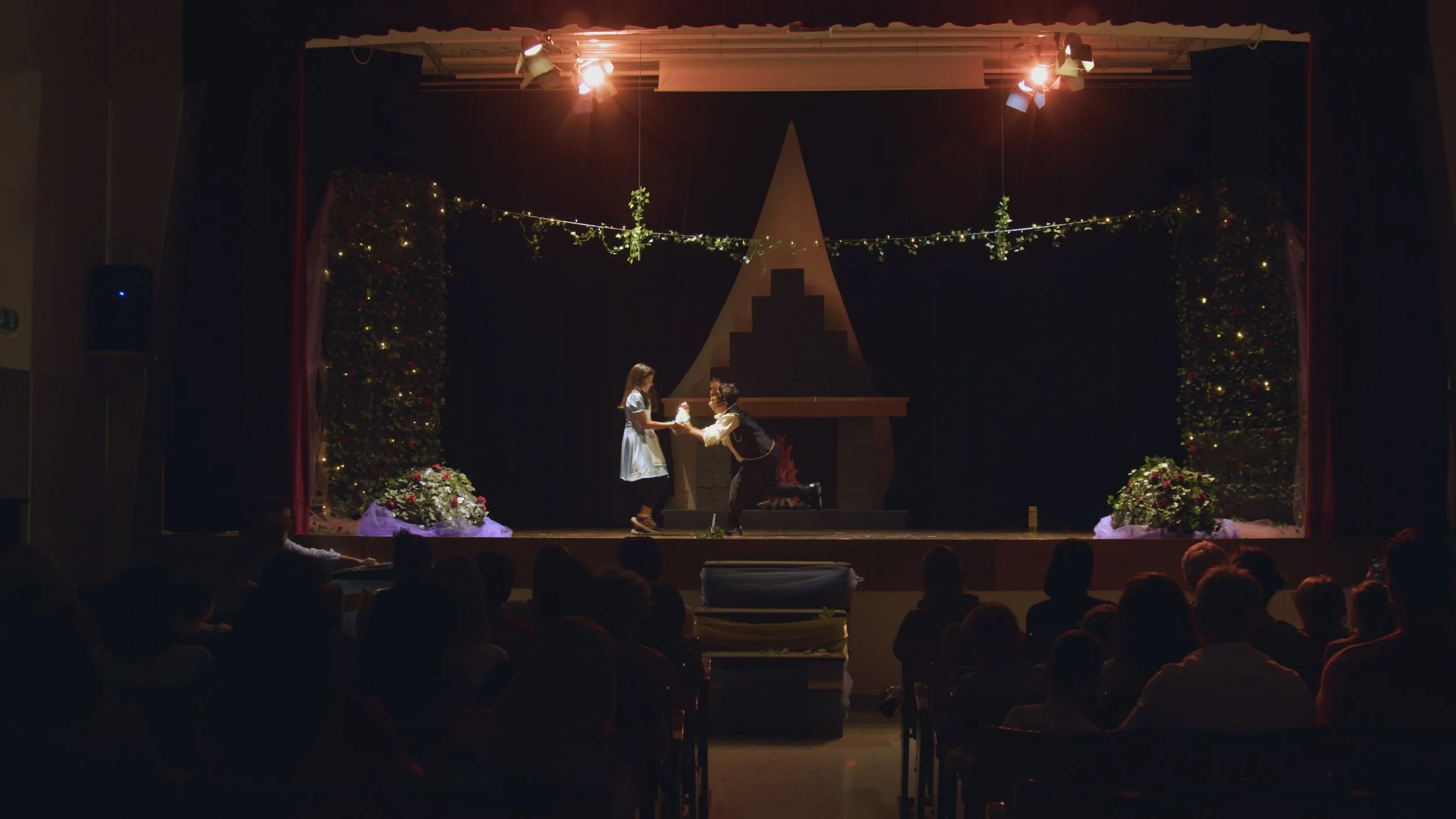 Scena teatrale con due attori vestiti in abiti storici, uno dà qualcosa all'altro, sul palco decorato con luci, piante e un camino, pubblico seduto in prima fila.