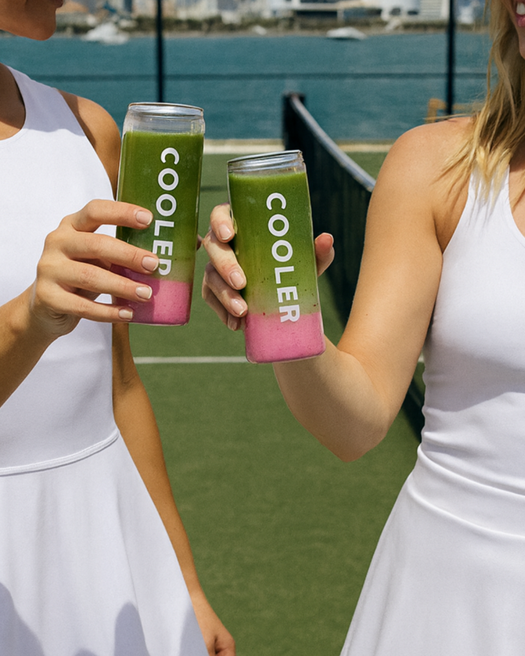 Two women in white tennis dresses holding glasses of colorful layered smoothies with green and pink sections, standing outdoors near a tennis court. Tennis event activations miami. padel event activations miami. On-site canning machine drinks