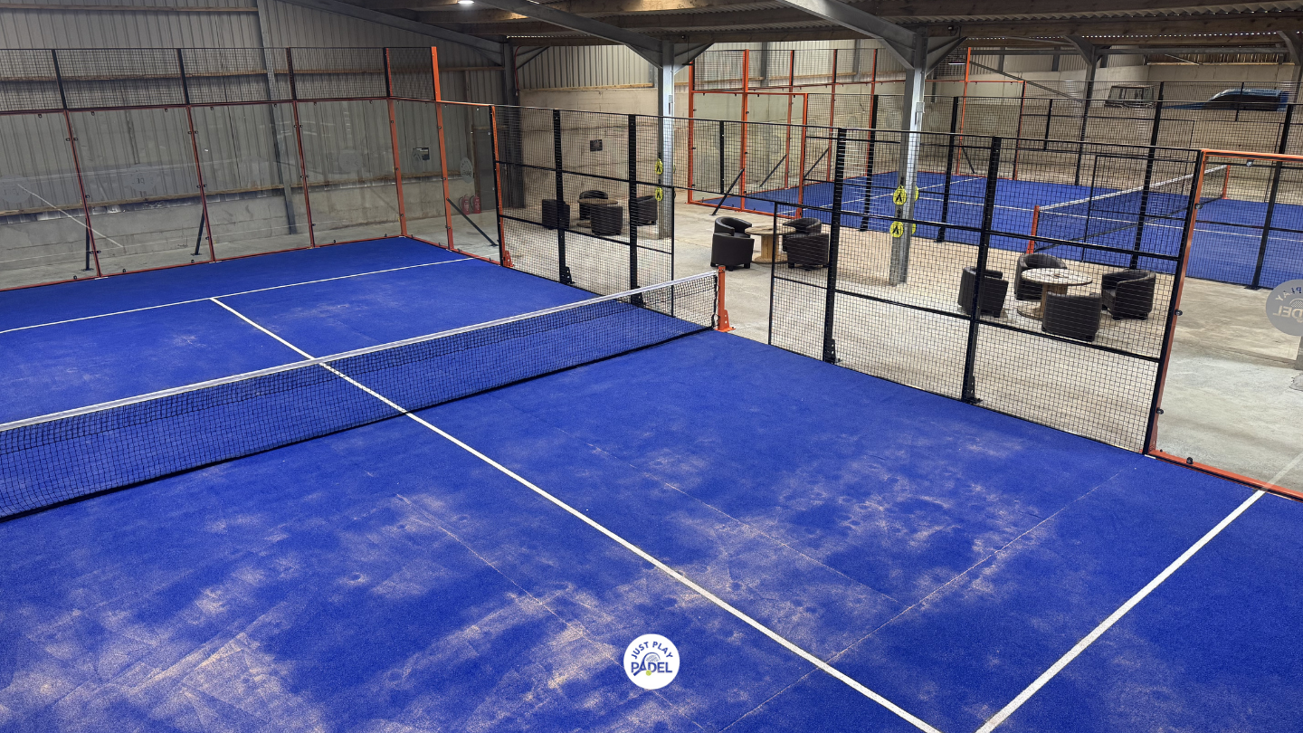 Indoor padel tennis court with blue surface and black net, surrounded by black fencing, with seating area and additional courts in the background.