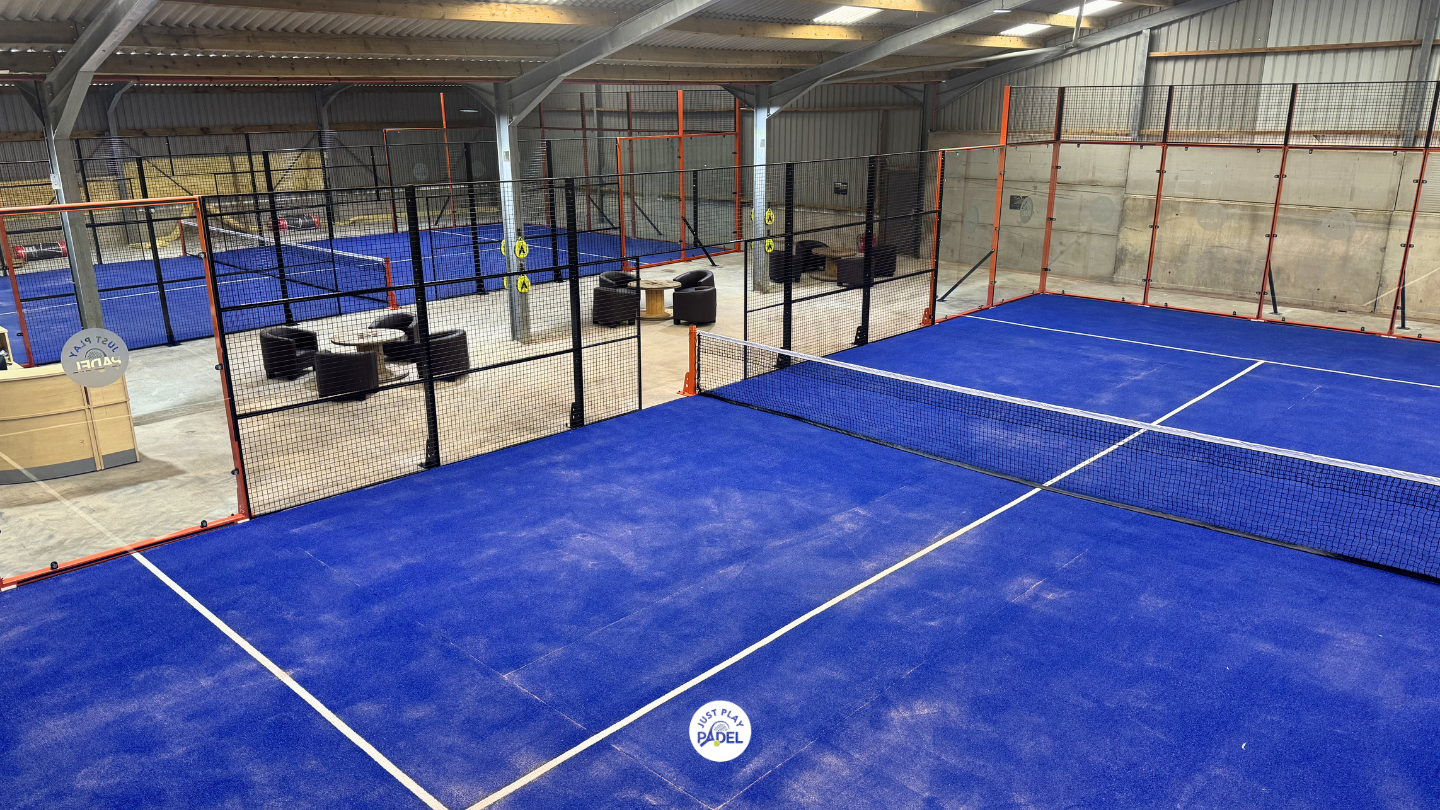Indoor sports facility with blue pickleball and tennis courts, surrounded by black and orange fencing, including seating and a bar area.