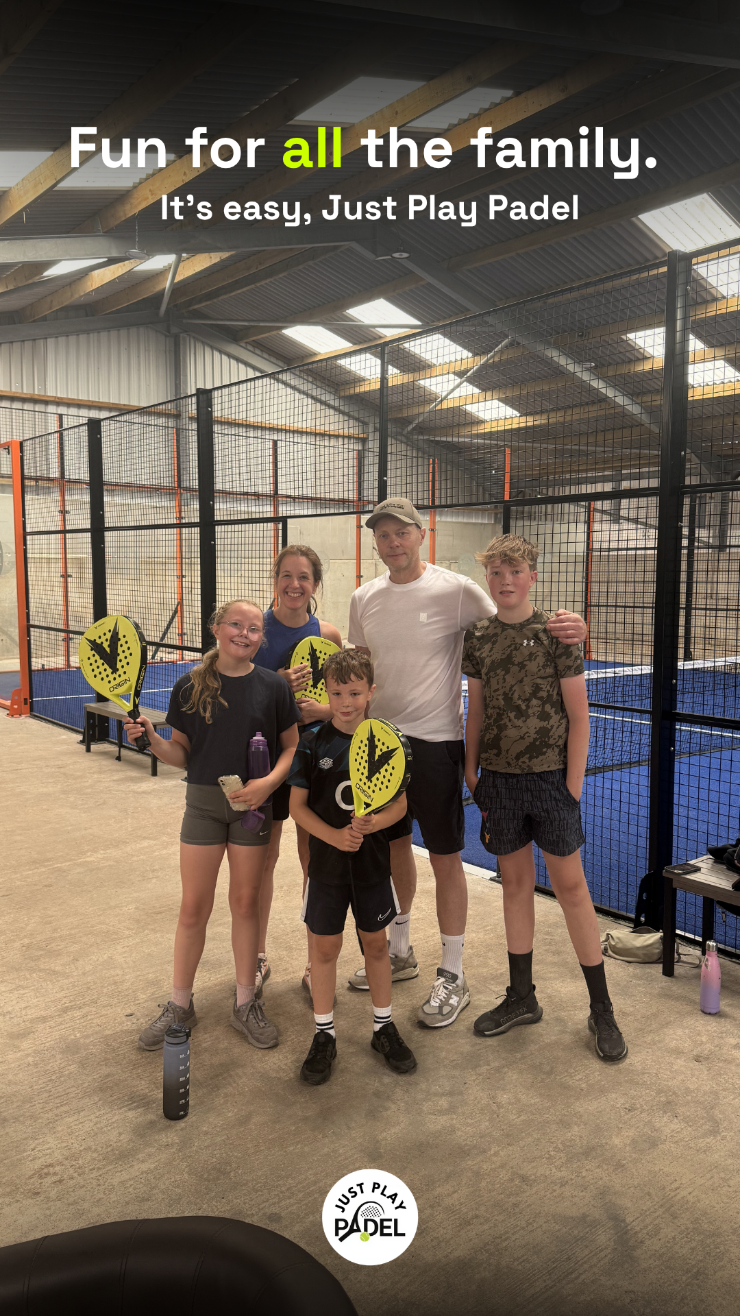 A group of five children and an adult man standing inside an indoor padel court, holding yellow padel rackets and smiling for the photo.