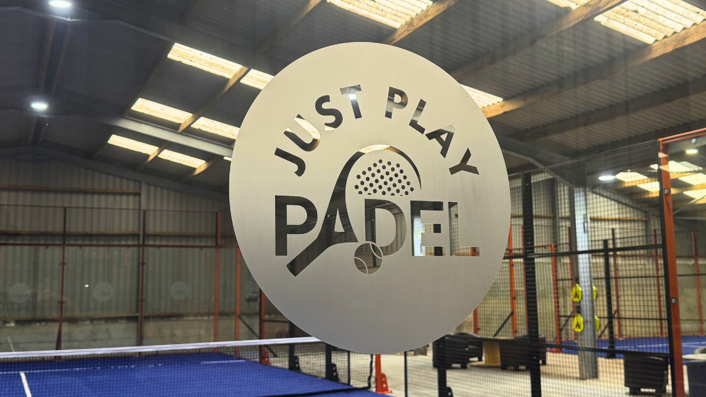 A round sign on a glass barrier with the words "Just Play Paddle" and a stylized paddle and ball logo, inside an indoor paddle tennis court.