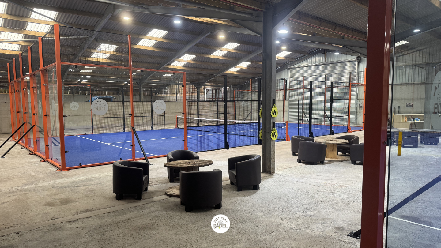 Indoor padel tennis court with seating area including black armchairs and small wooden tables.