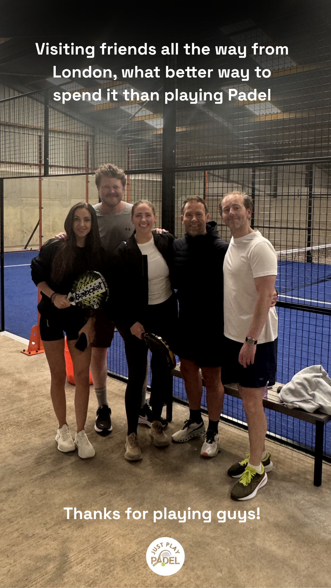 Group of five people standing inside an indoor padel court, smiling and holding padel rackets. There are tennis or padel courts in the background, with one court visible with blue flooring. The group appears to be enjoying a game or celebrating after playing padel tennis.
