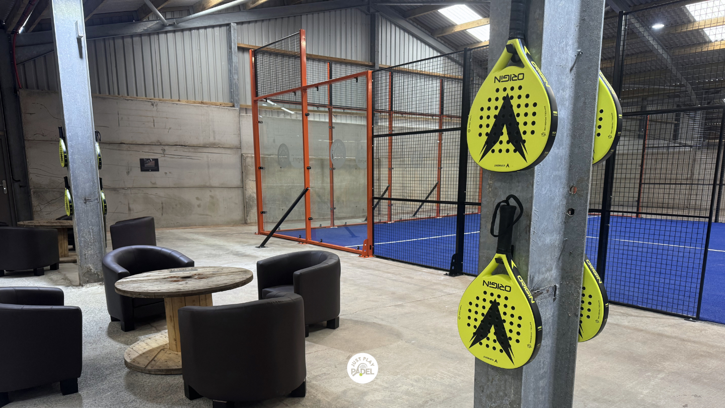 Indoor padel court with blue playing surface, surrounded by black metal fencing. Four black armchairs around a round wooden table in the lounge area. Several yellow padel rackets hung on a concrete pillar.