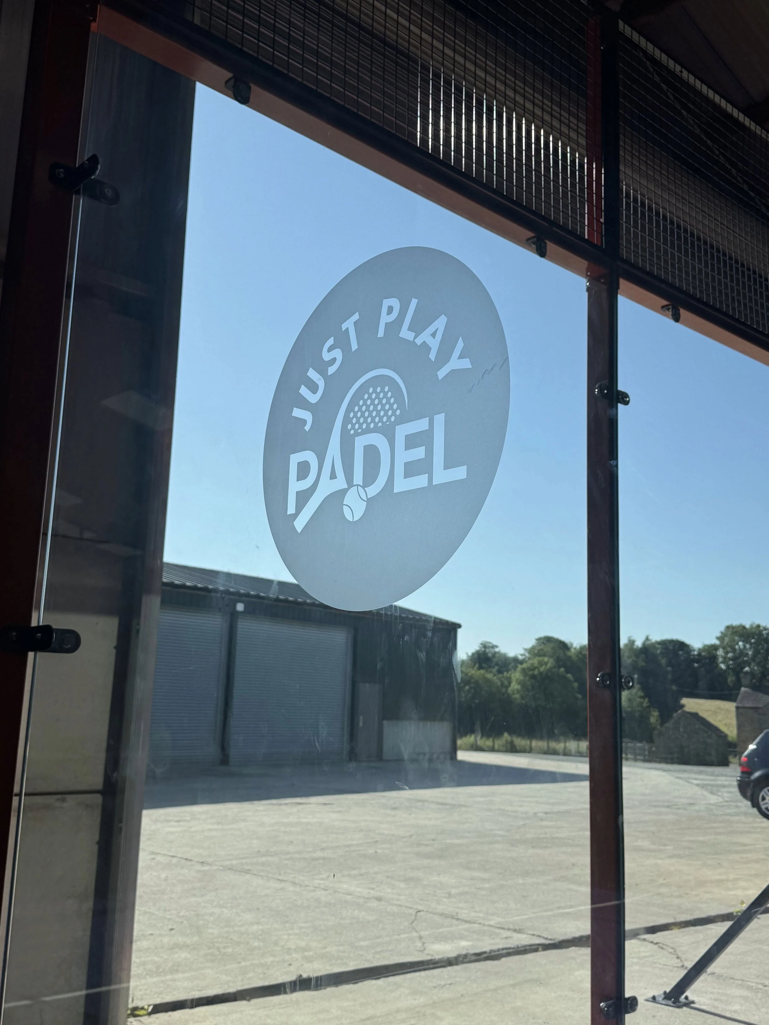 A clear glass window with a logo that reads "Just Play Padel" and a small paddleball illustration. Outside, a parking lot with a building, trees, and a parked car is visible under a blue sky.