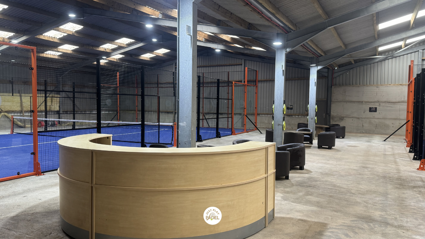 Indoor padel tennis facility with multiple courts separated by black and orange fences, a reception desk at the front, and seating area with black chairs and stools.