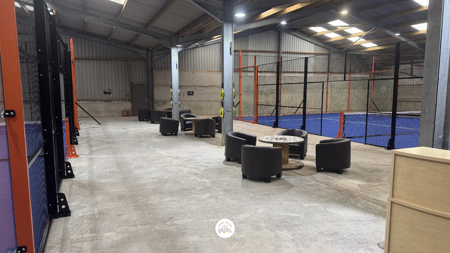 Indoor sports facility with a padel tennis court enclosed by a black and orange net. Seating area with black chairs and a round table, concrete floor, and a metal roof.