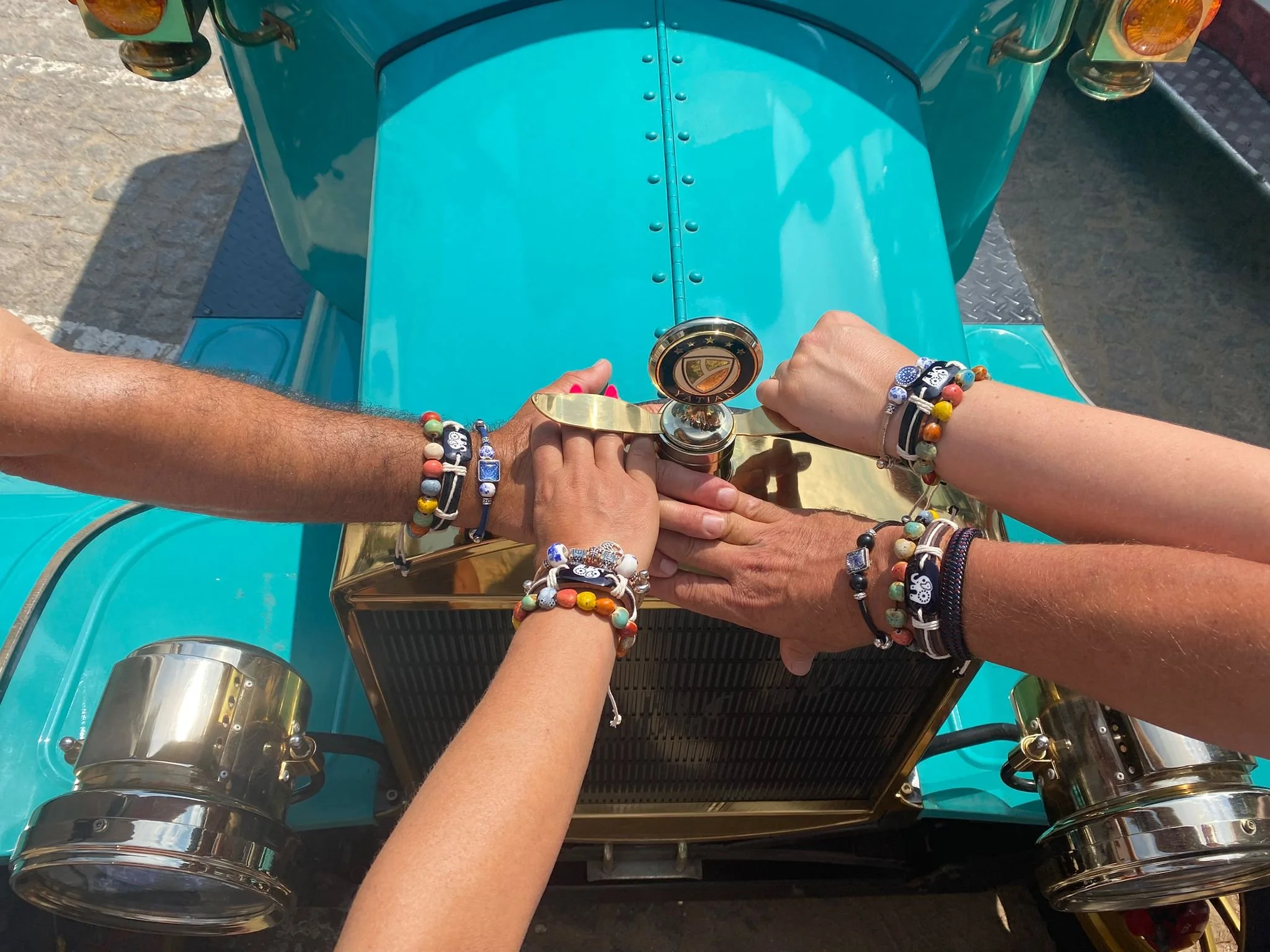 Mãos de várias pessoas colocadas uma sobre a outra, segurando a chave de um carro antigo de cor azul com detalhes em dourado, em cima do capô do carro.