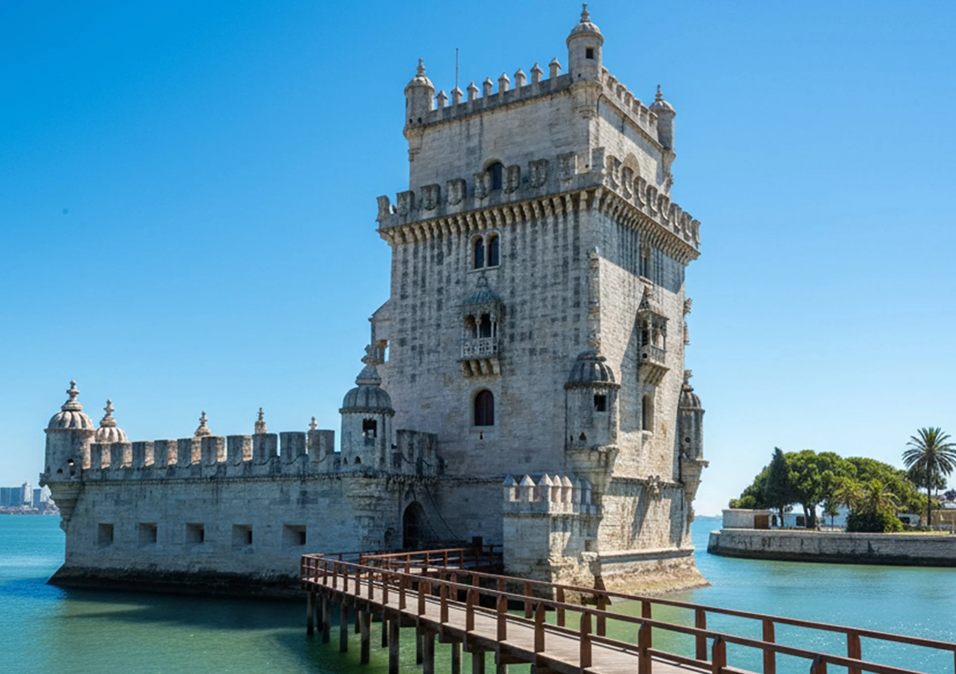 Castelo de Belém, Lisboa, Portugal, visto ao lado do rio, com céu azul claro ao fundo, árvores verdes na margem, e ponte de madeira na frente.