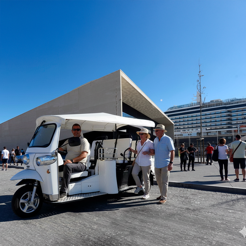 Pessoas usando um carrinho de golfe na área externa de um edifício moderno, ao lado de um grande navio de cruzeiro.