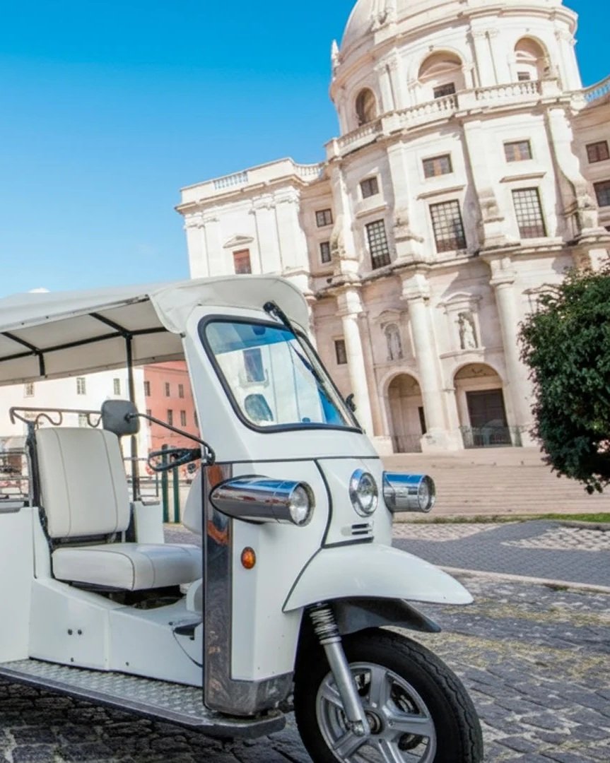 Uma motoneta de três rodas branca estacionada na rua com um edifício histórico de arquitetura clássica ao fundo