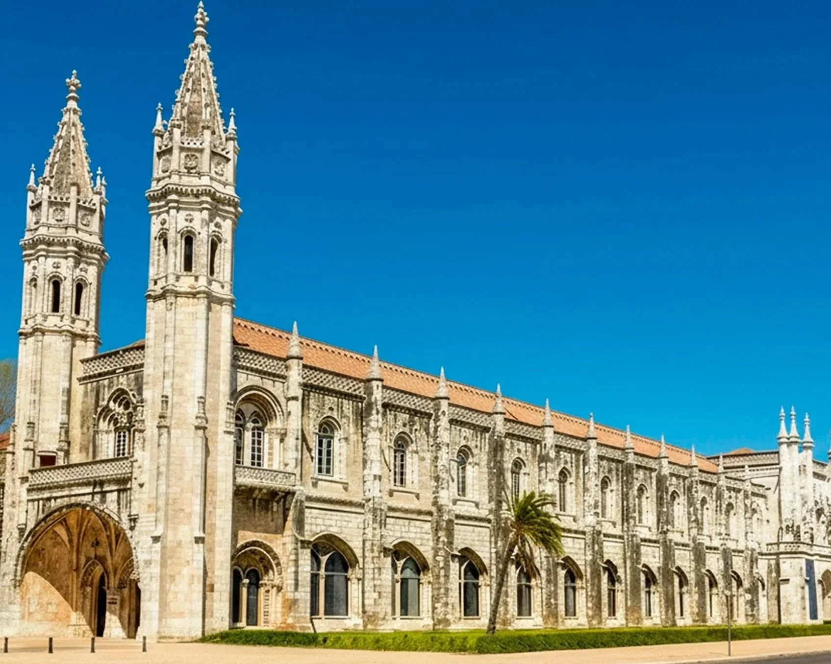 Igreja histórica com torres góticas e detalhes arquitetônicos de pedra, com céu azul ao fundo, e uma árvore à frente.