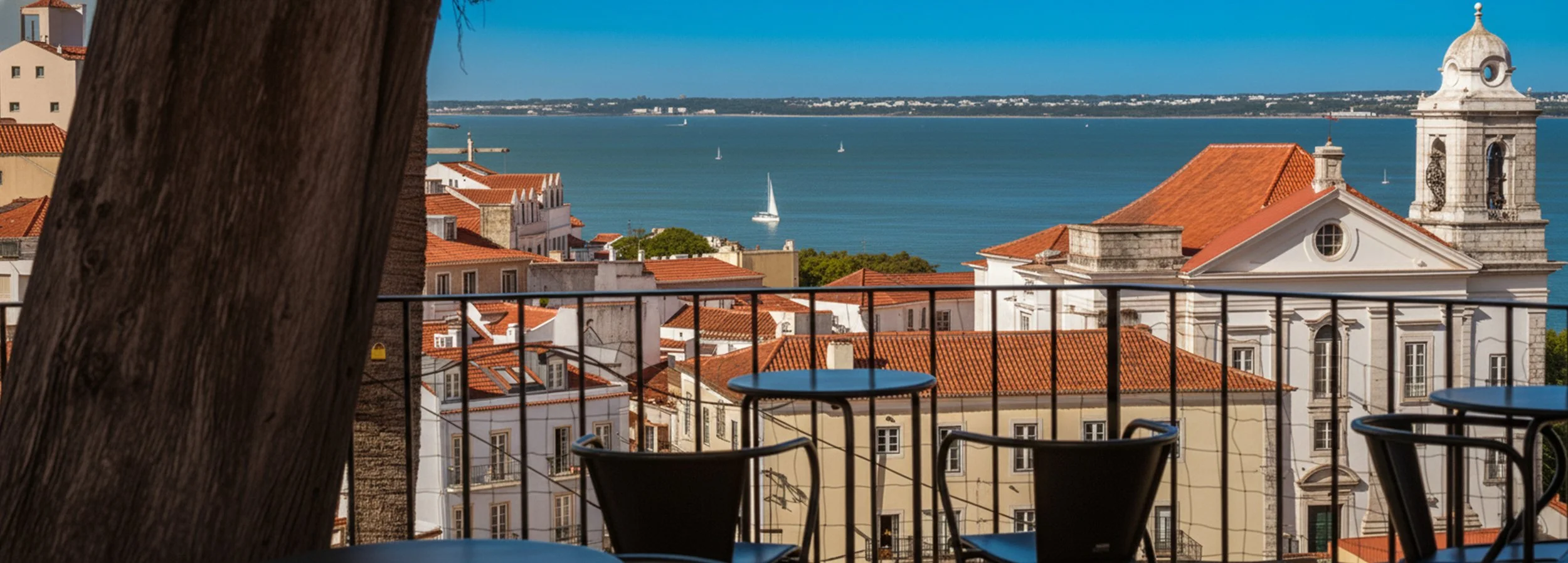 Vista de uma cidade costeira com prédios de telhados vermelhos, uma igreja com cúpula e vista para o mar com veleiros ao fundo, vista de uma varanda com cadeiras e mesas de vidro, uma árvore na esquerda parcialmente visível.