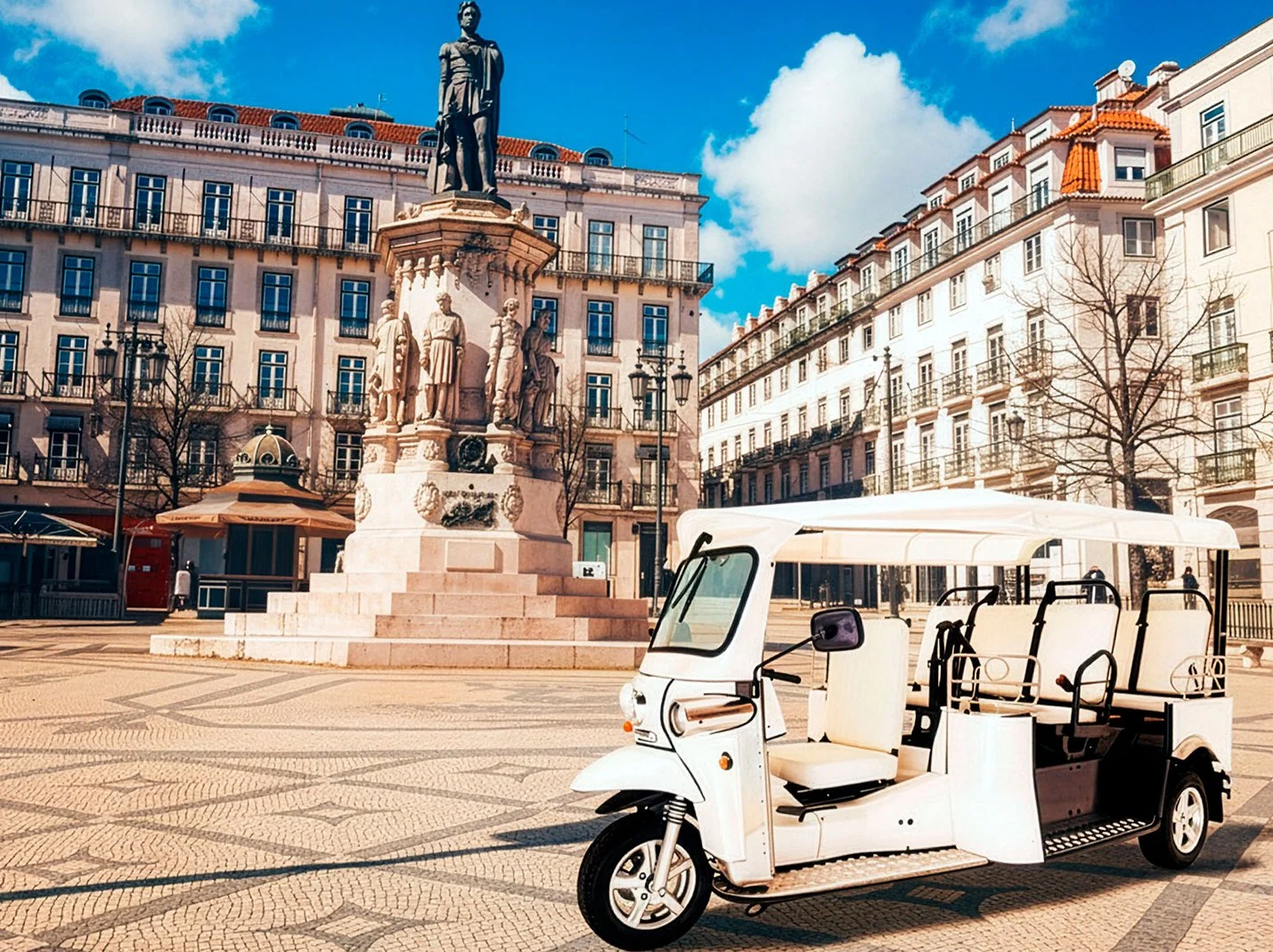 Praça com uma estátua de um homem no centro, cercada por edifícios de vários andares com janelas e varandas, e um carrinho elétrico no primeiro plano. Céu azul com algumas nuvens.