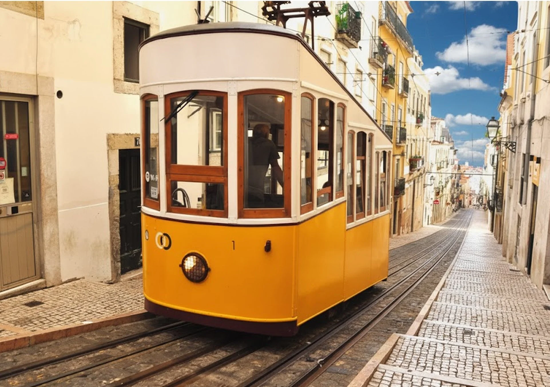 Bondinho amarelo antigo passando por uma rua de paralelepípedos em uma cidade com prédios coloridos de altura baixa e céu azul com nuvens.