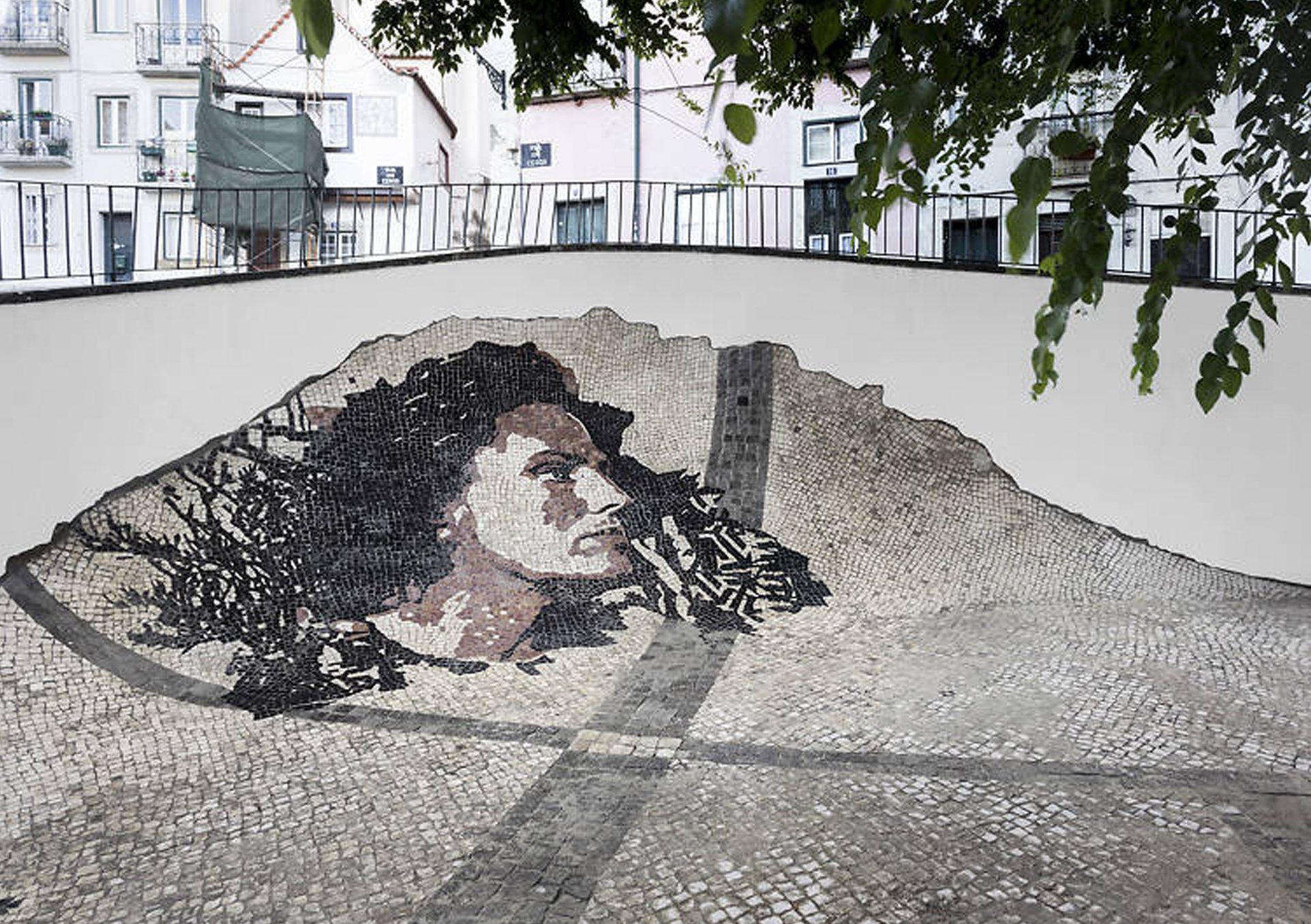 Mosaico de rua com retrato de uma mulher com cabelo ondulado e expressão séria, feito com tesselas de cores variadas, localizado em uma área ao ar livre cercada por prédios.