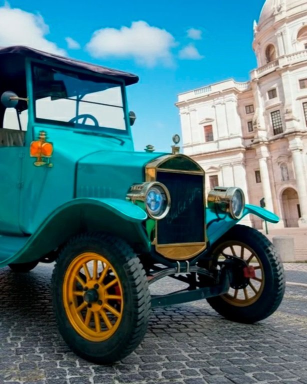 Automóvel antigo de cor verde com detalhes dourados, rodas amarelas, em frente a uma arquitetura histórica de pedra, sob céu azul com algumas nuvens.