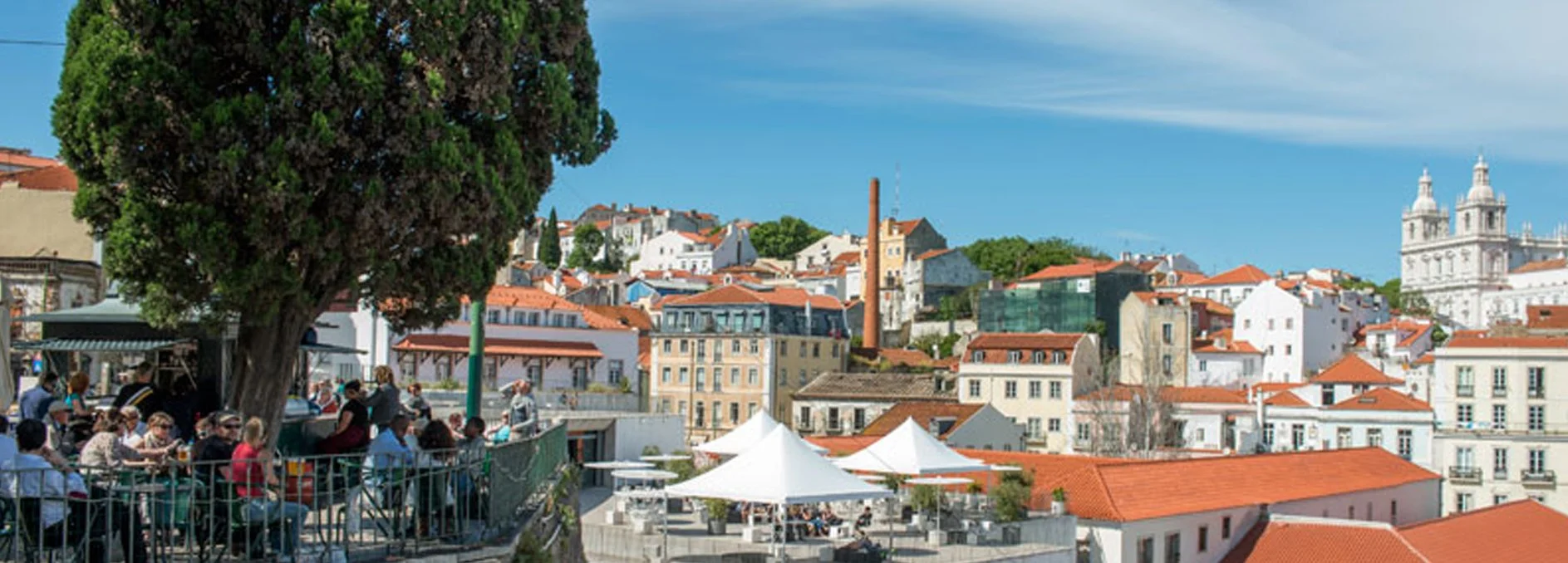 Vista de uma cidade com prédios de telhado vermelho, uma grande árvore e pessoas sentadas em uma praça com guarda-corpo de ferro, céu azul com algumas nuvens.