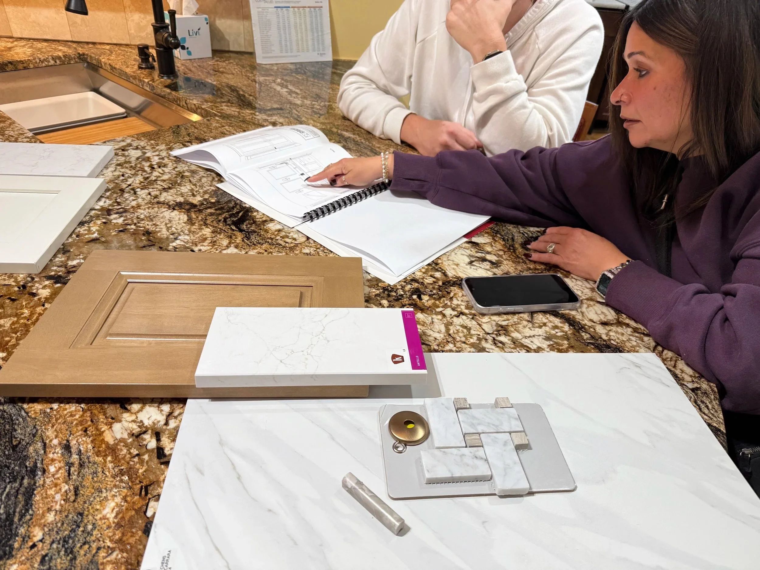 Two women at a kitchen showroom discussing cabinet designs, with color and material samples on the counter.