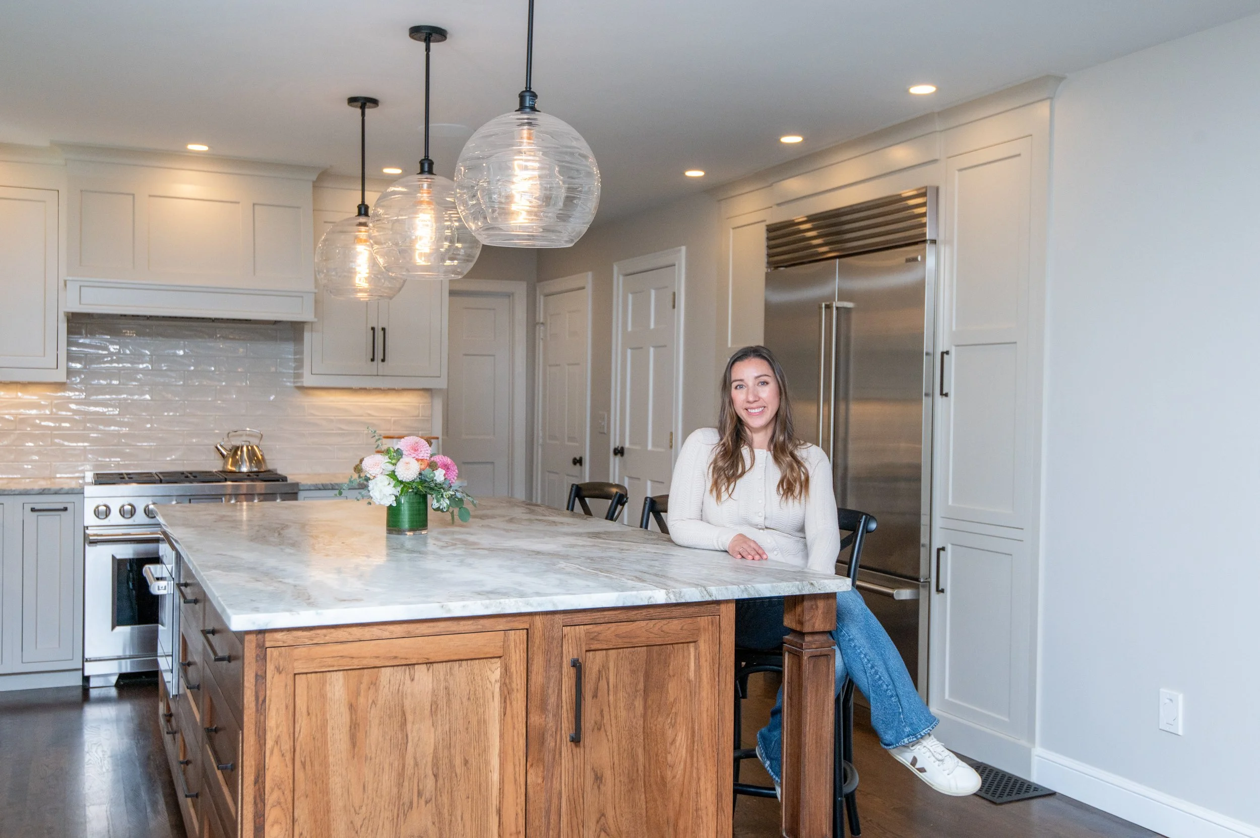 A young woman sitting on a stool in a modern kitchen with white cabinets, stainless steel appliances, and a large marble island with a flower arrangement. She is smiling, wearing a white sweater and blue jeans.