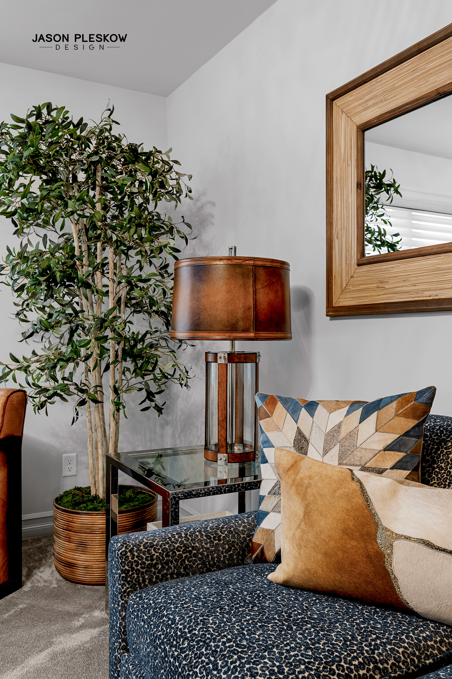 Living room corner with a patterned sofa, decorative pillows, a tall potted plant, a glass side table with a leather and wood table lamp, a wooden framed mirror on the wall, and a white wall with text 'JASON PLESKOW DESIGN' in the top left corner.