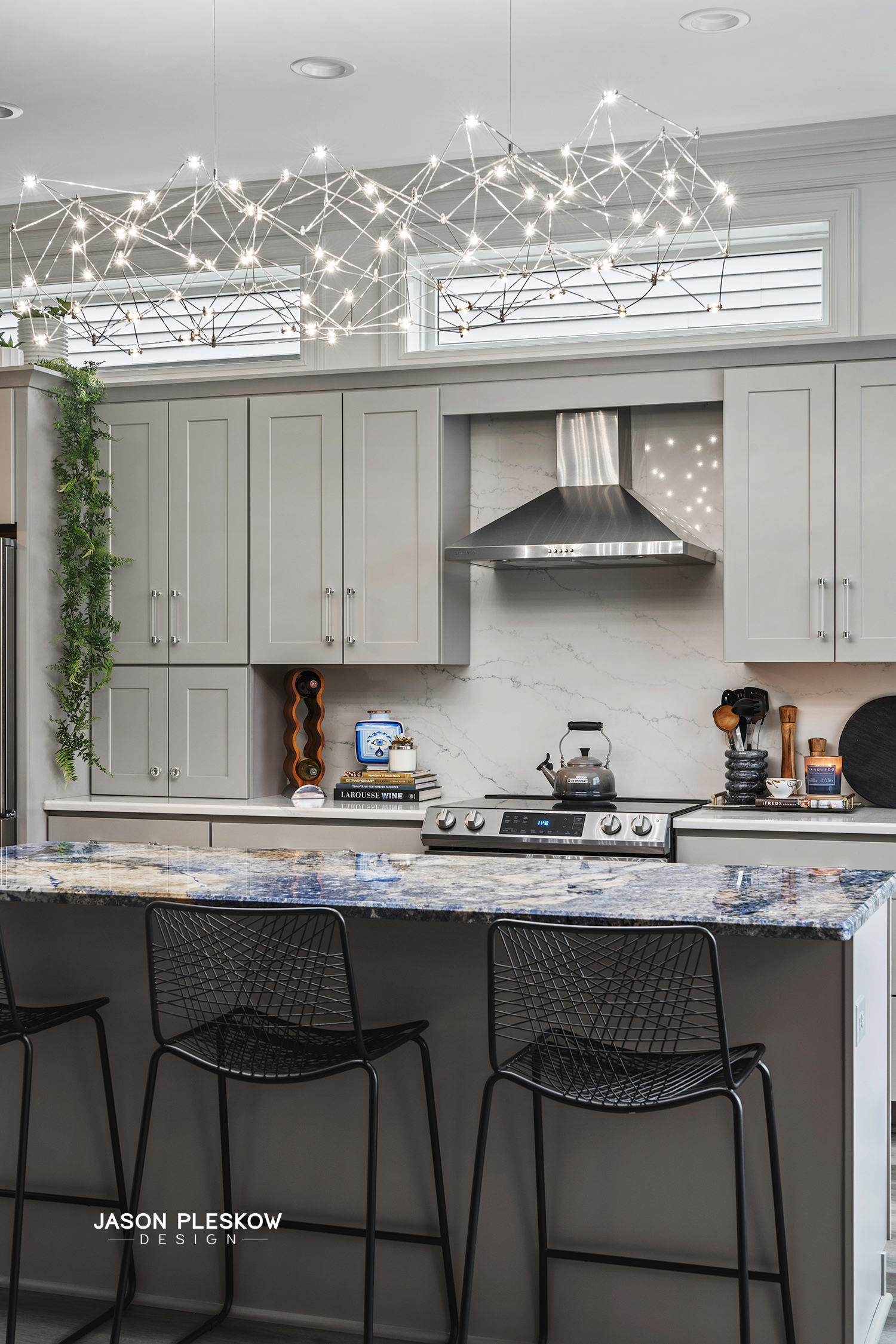 Modern kitchen with gray cabinetry, marble backsplash, stainless steel range hood, black wire bar stools, and geometric LED light fixtures.