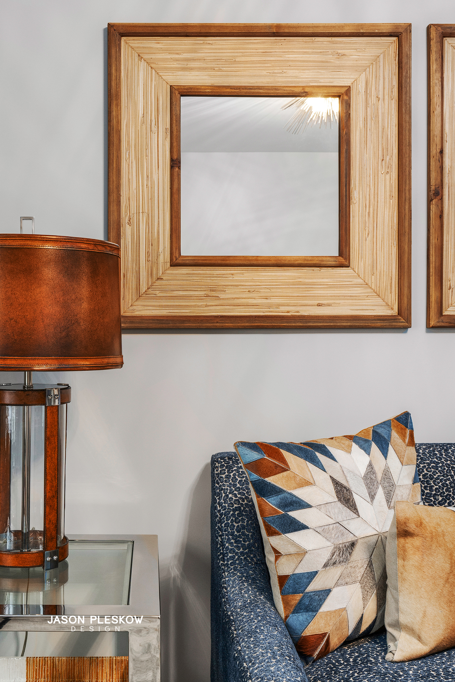 Interior living room decor featuring a square wooden mirror on a light-colored wall, a table lamp with a brown shade on a side table, and a tufted blue armchair with decorative pillows, one with a geometric pattern with shades of blue, brown, and bei