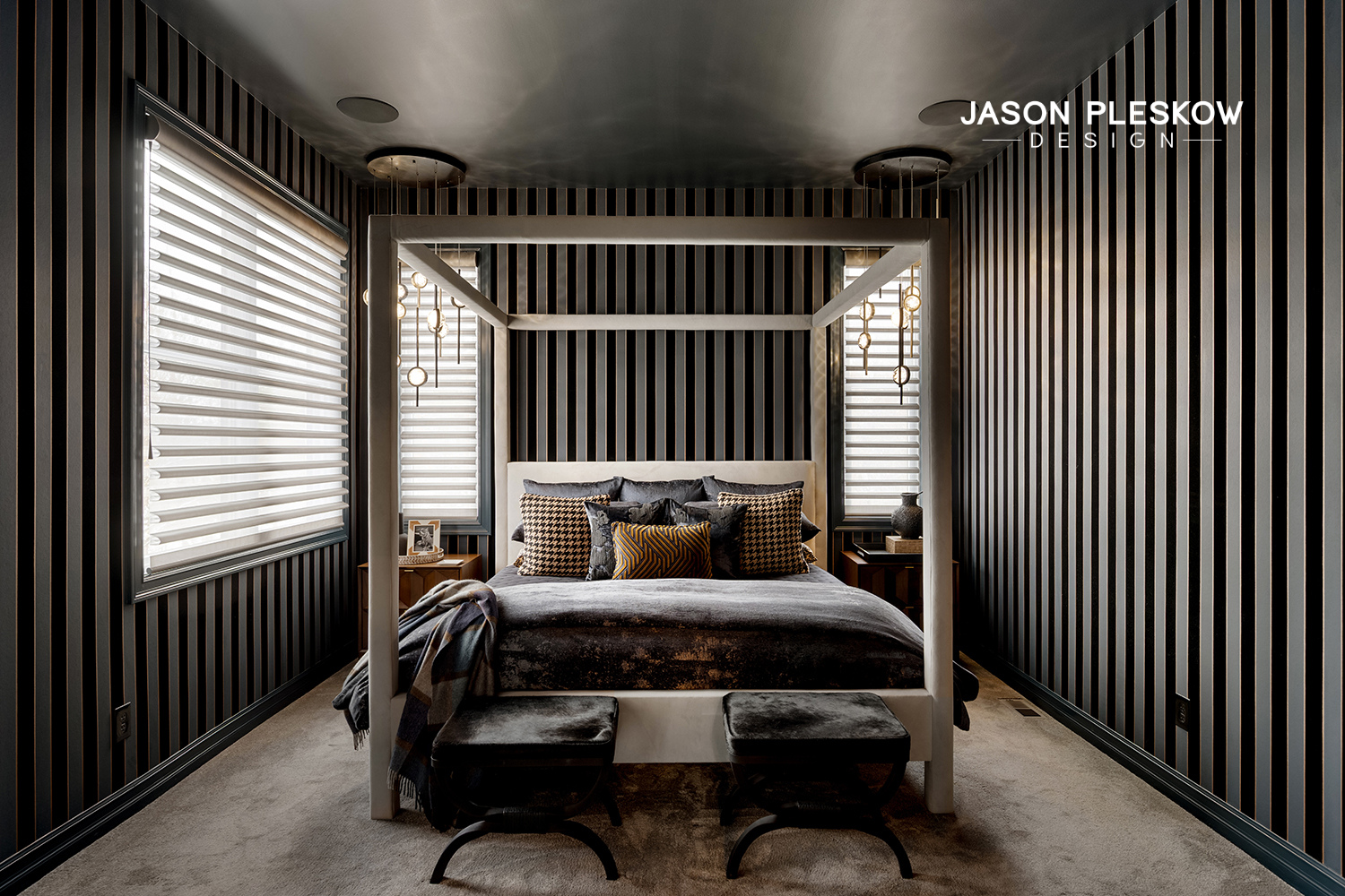 A modern bedroom with black vertical striped walls, a white four-poster bed with dark bedding, and a variety of patterned pillows. Two nightstands with decorative items are on either side of the bed. There are large windows with white blinds, modern 