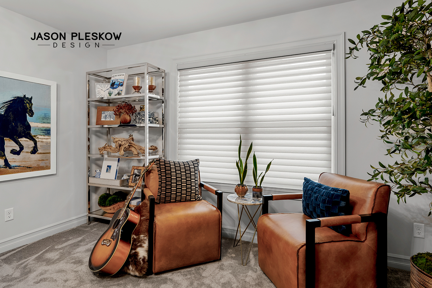 A cozy living room corner with two brown leather armchairs, decorative pillows, and a guitar resting on one chair. A small round marble table with potted plants is between the chairs. There is a white window with closed blinds, a tall shelving unit w