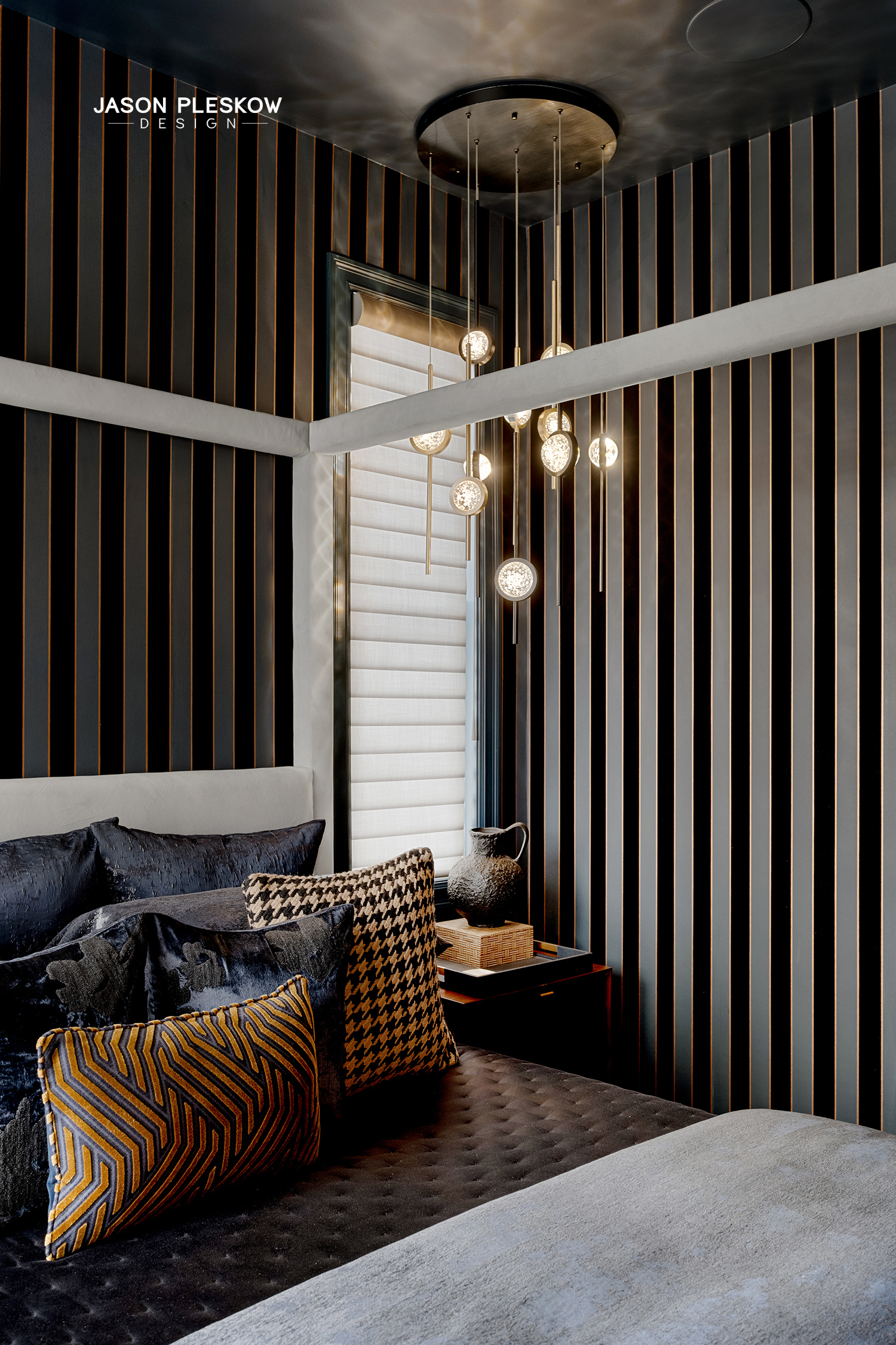 Interior view of a bedroom with black and gold vertical striped wall, modern chandelier with round glass bulbs, window with blinds, black and patterned pillows on a bed, and a bedside table with a decorative vase and tray.