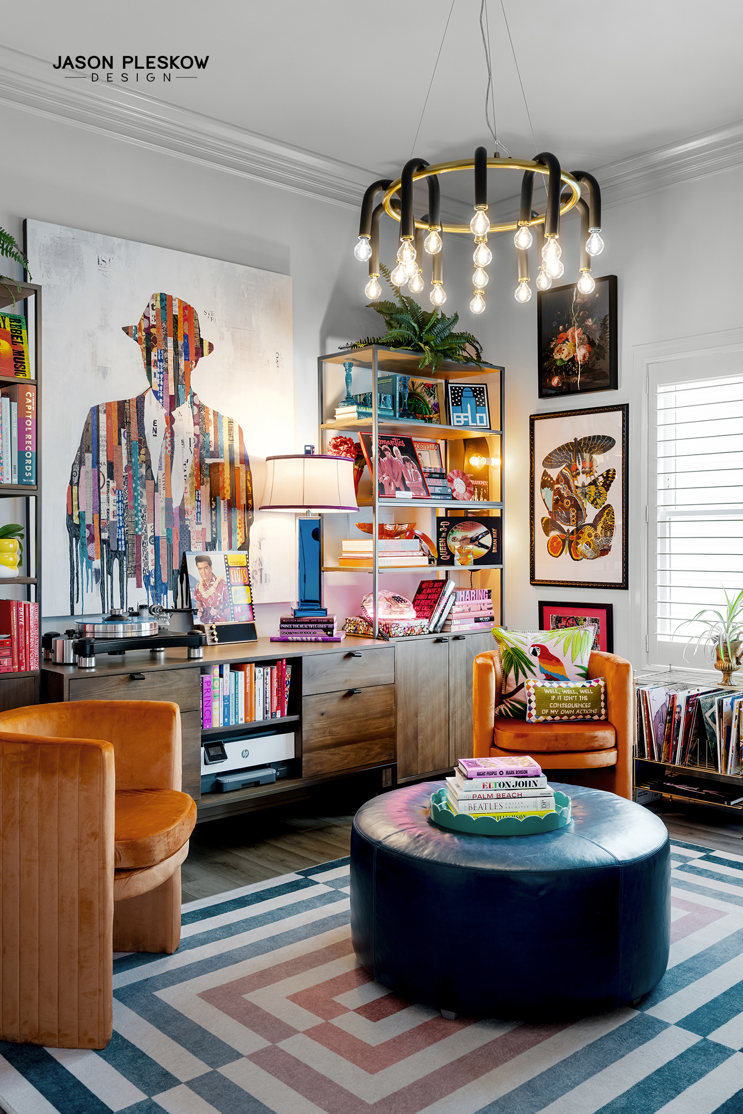 Colorful living room with orange armchair, black ottoman, and wooden shelving unit decorated with books, art, and plants. Large abstract portrait of a man on the wall, and a modern chandelier hanging from the ceiling.