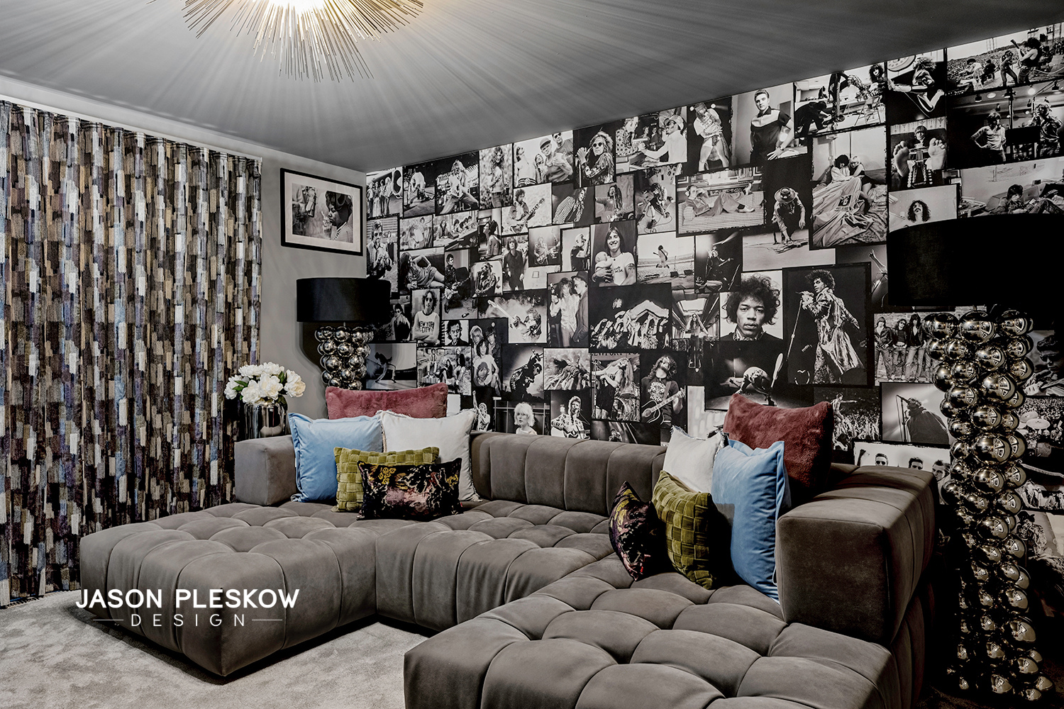 Living room with a large gray sectional sofa decorated with colorful pillows, a wall covered with black and white photos, a side lamp with a chrome ball base, and a floral arrangement on a side table. The room has patterned curtains and a textured ce