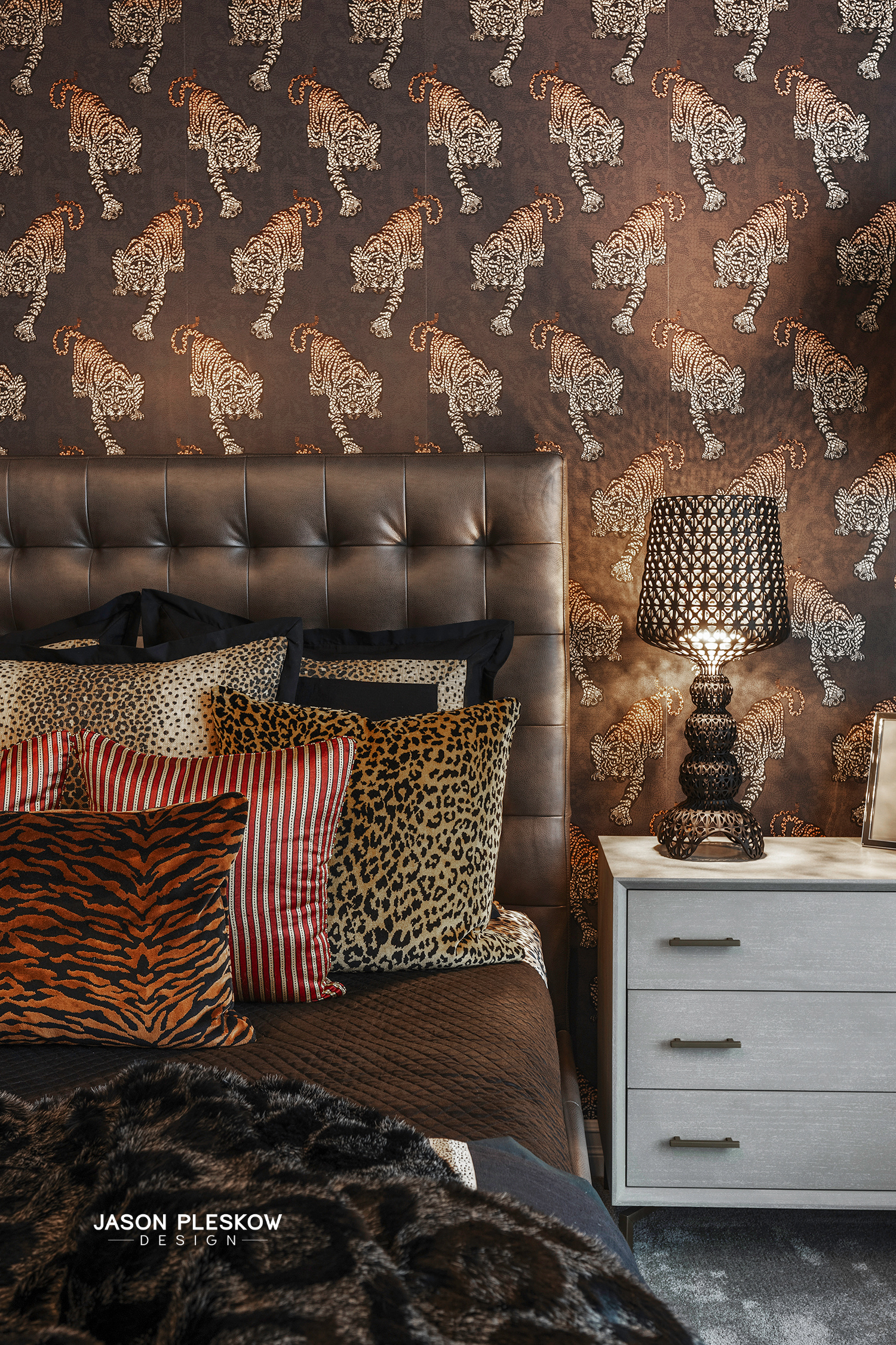 A bedroom with animal print decor and tiger wallpaper featuring white tigers. Multiple animal print pillows are on the bed, including leopard, zebra, and tiger patterns. A modern, black table lamp with a geometric design is on a white nightstand besi