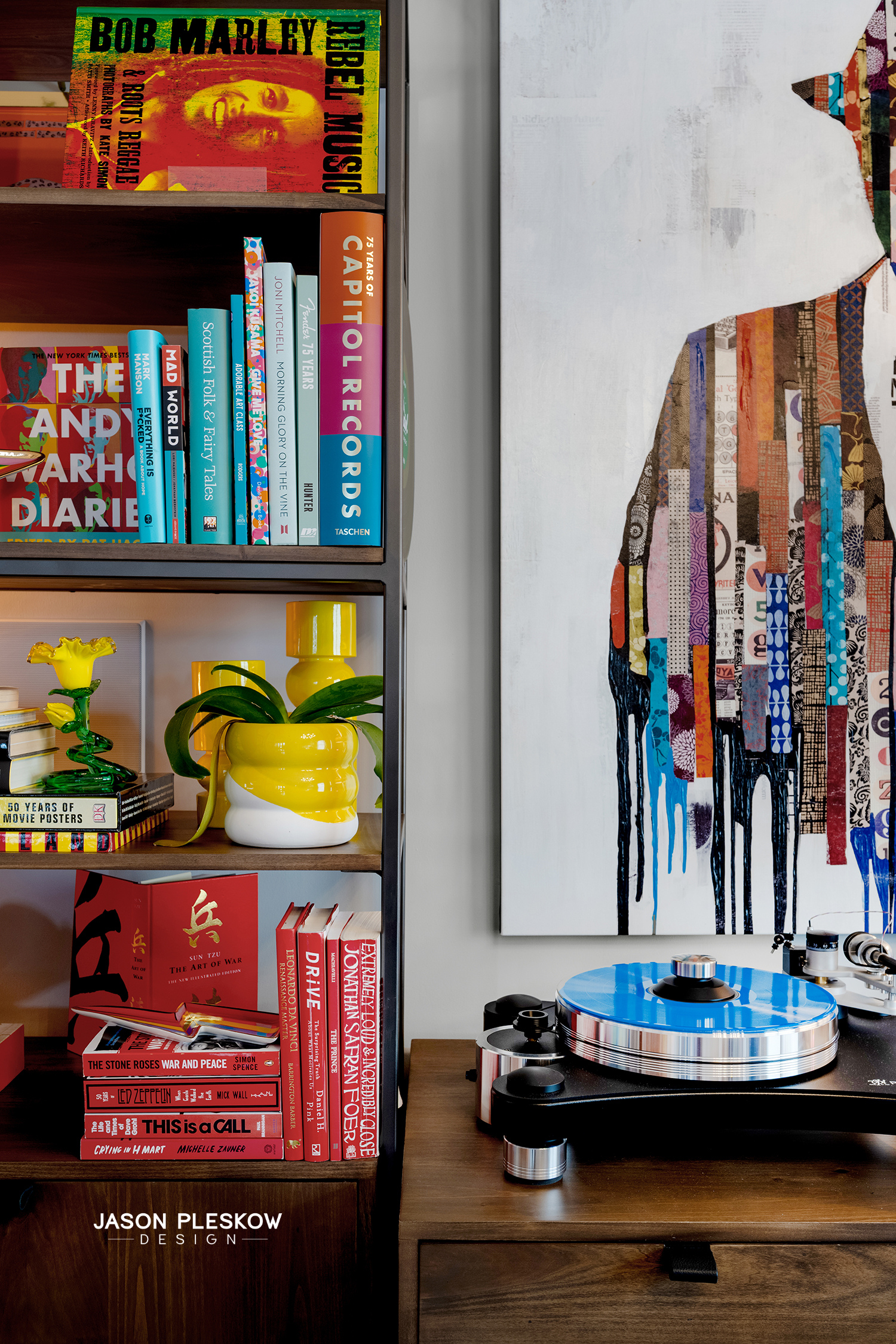 A bookshelf with colorful books, yellow and white vases, a yellow flower sculpture, and a record player on a wooden table, with abstract wall art featuring colorful vertical strips and paint drips.