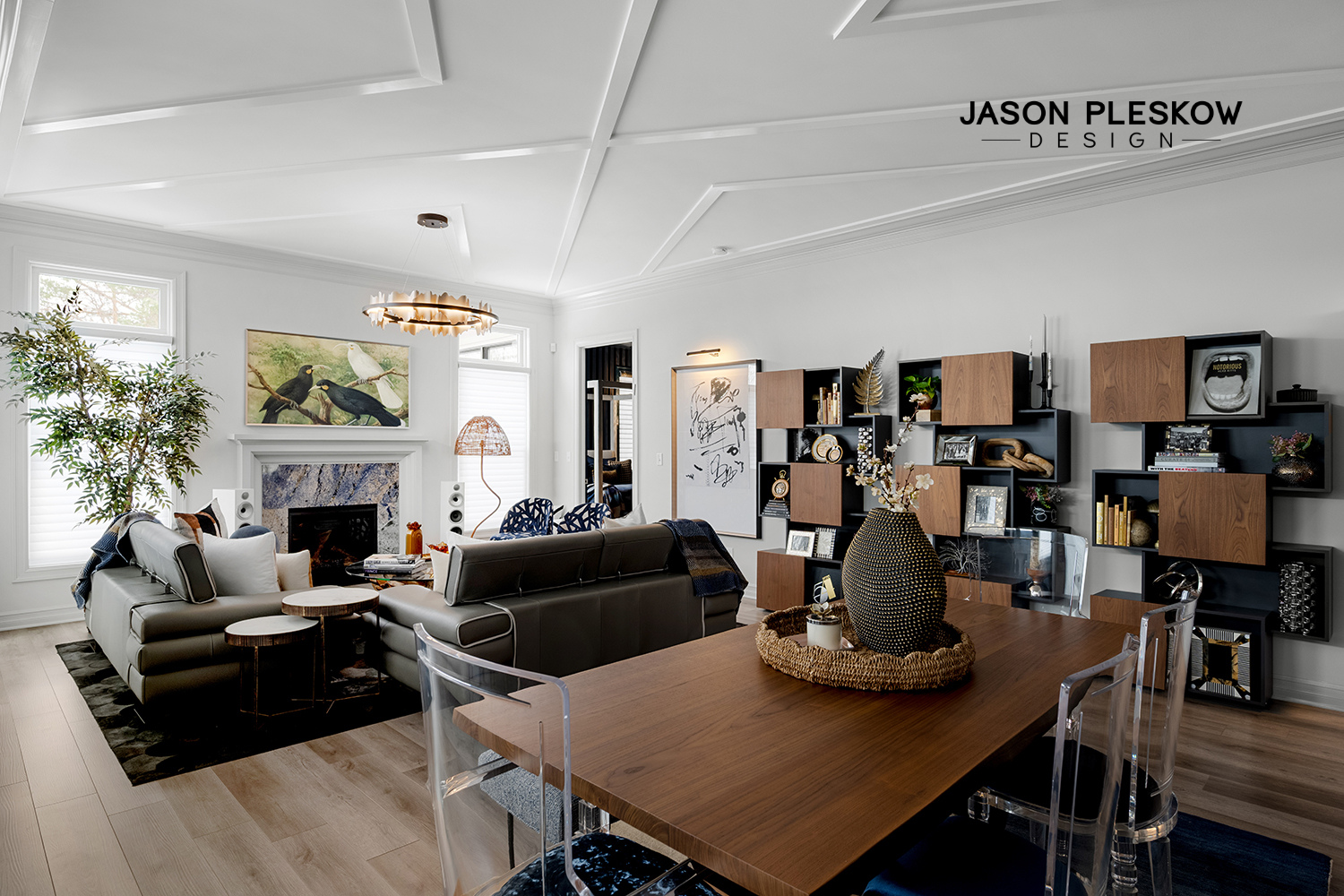 Modern living room with white walls, a fireplace, and large windows. Furnished with a black leather sectional sofa, white pillows, an abstract paintings on the wall, a decorative vase on the dining table, and a wall-mounted shelving unit with books a