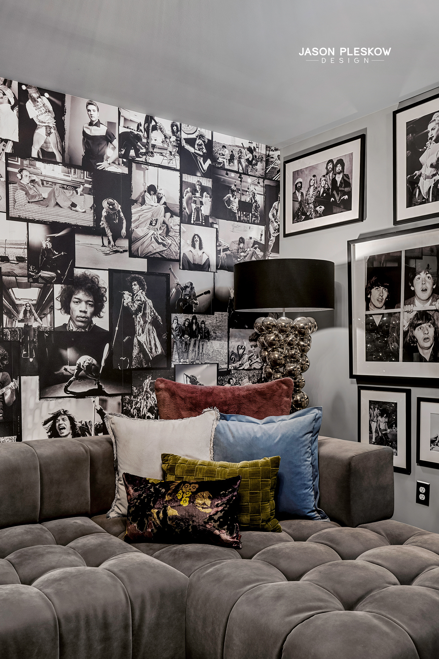 A living room corner with a grey tufted sofa adorned with various colorful pillows, including white, blue, red, green, and black with floral pattern. Behind the sofa, there is a black and white photo wall featuring numerous framed photos of famous mu